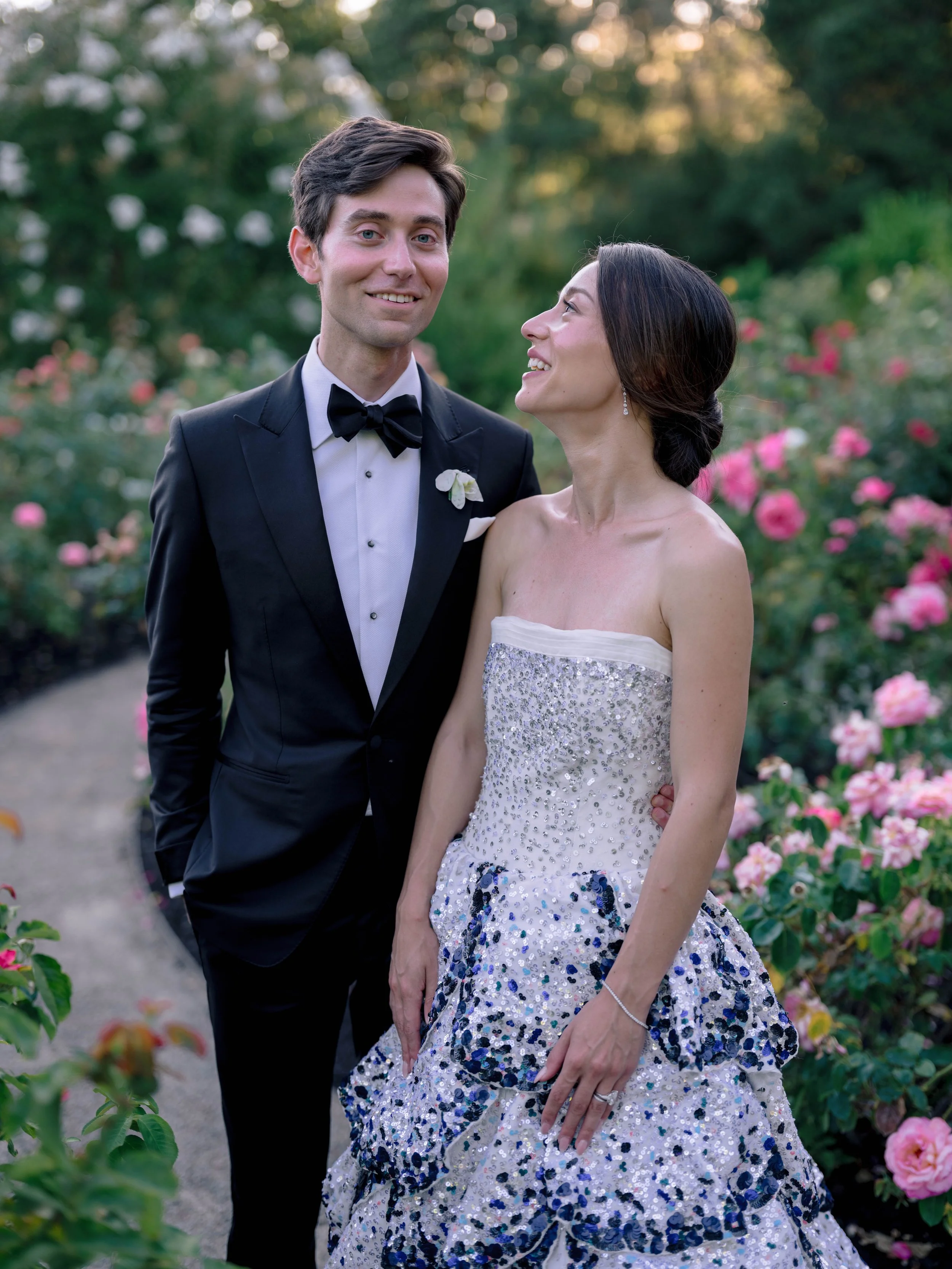 A young man and woman in formal attire posing outdoors in a garden with pink and white flowers, smiling at each other.