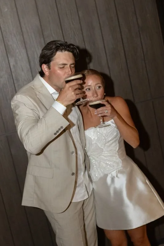 A man in a beige suit and a woman in a white dress are holding drinks and appear to be at a party or celebration, standing against a wooden-paneled wall.