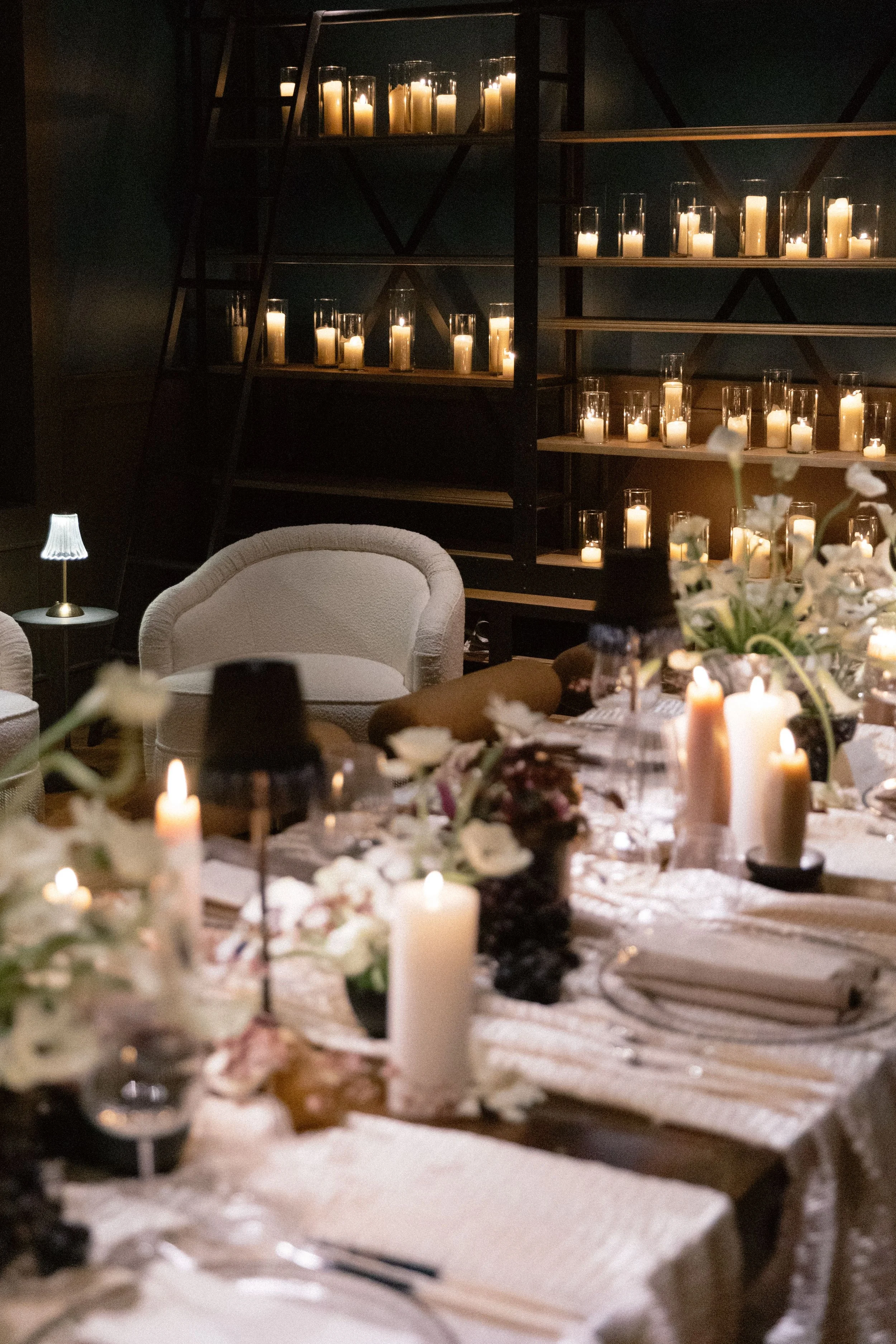 A table decorated with candles, flowers, and other decorations, set for a meal or celebration. In the background, a bookshelf with lit candles adds to the cozy ambiance.