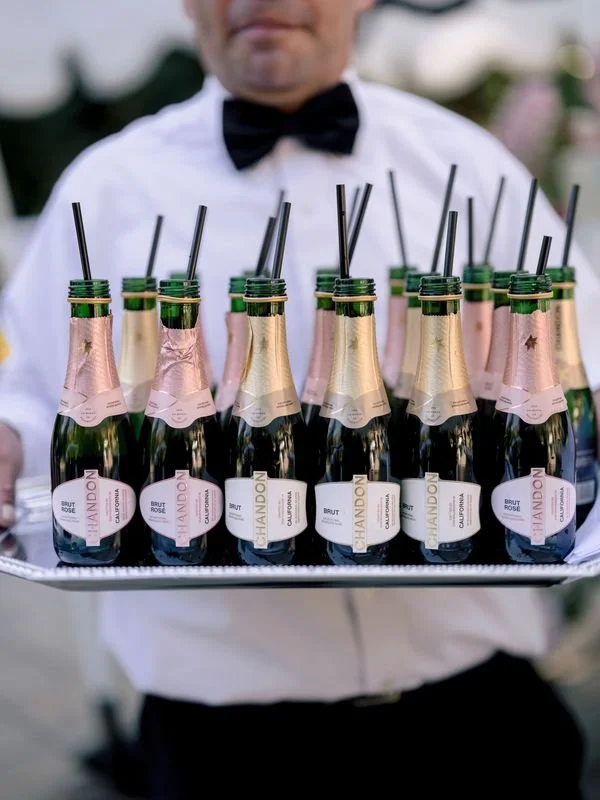 A waiter holding a tray with several mini bottles of sparkling wine decorated with gold and white foil, each with a black straw, at a festive event.
