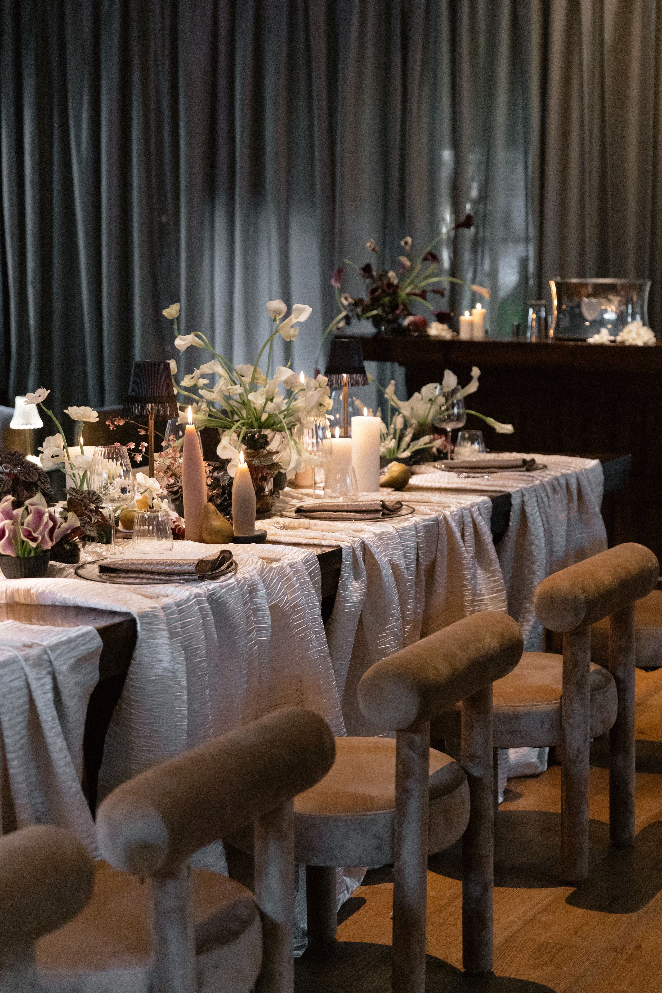 A dining table with white textured tablecloth, decorated with white flowers, candles, and glassware, set for a formal meal. In the background, a dark wooden sideboard and a dark curtain can be seen.