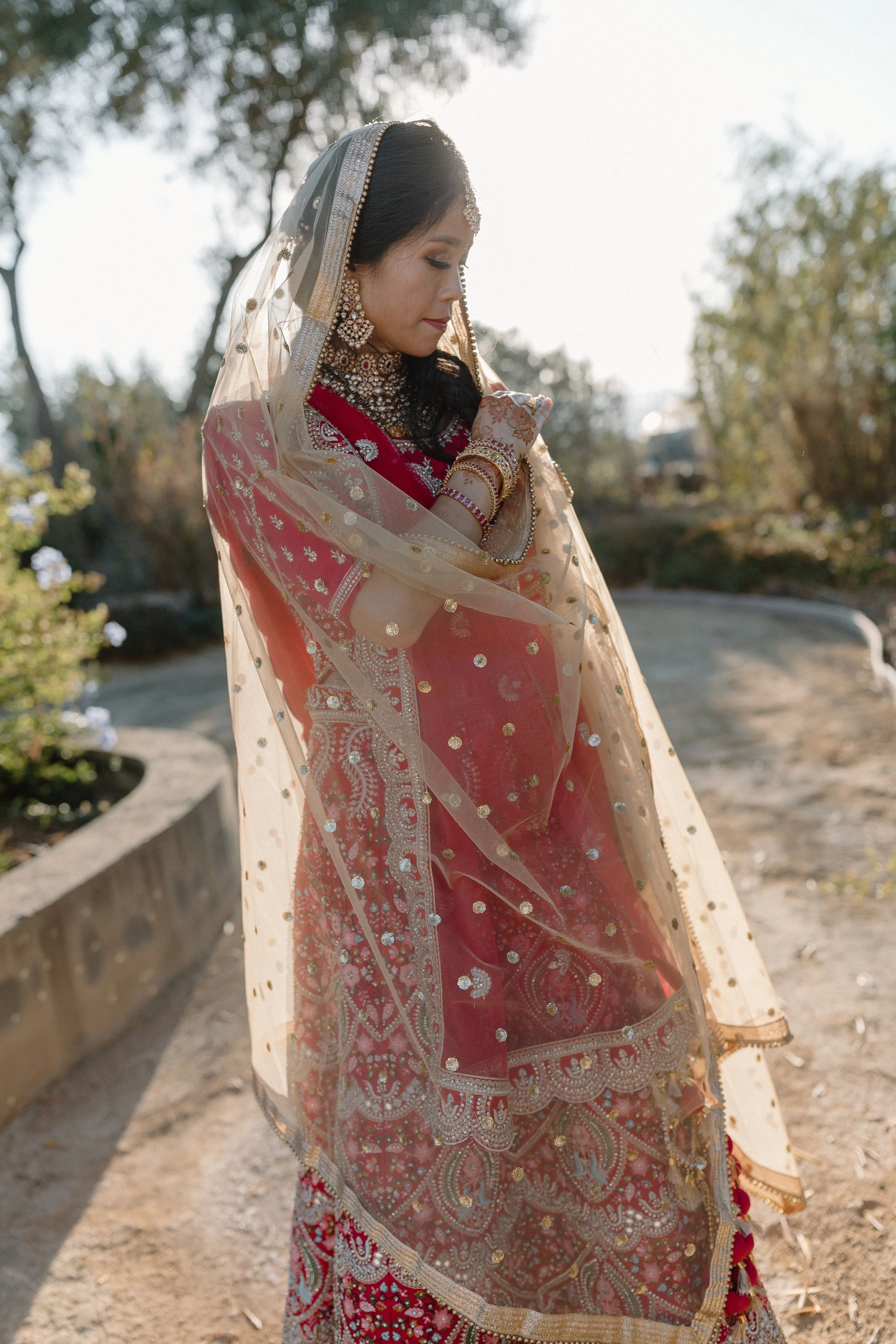 A woman in traditional Indian attire standing outdoors, looking down with her hands folded at her chest. She is wearing a red and gold outfit with intricate jewelry, including earrings, a necklace, and bangles. A sheer dupatta with gold accents drape