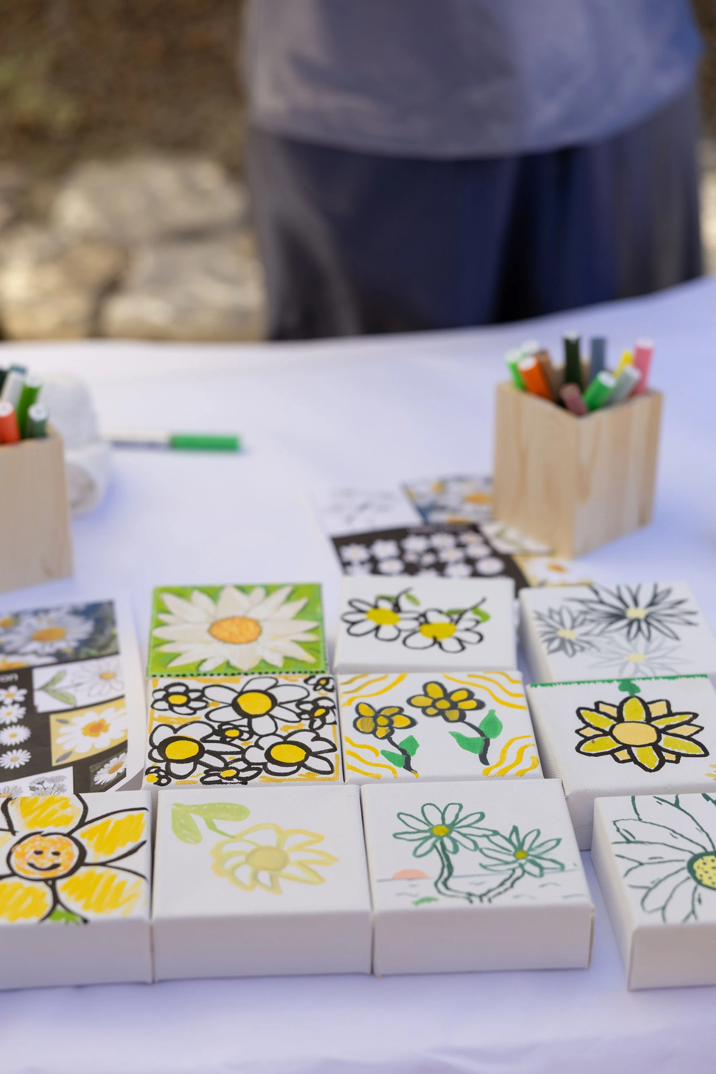 Colorful handmade tile art featuring various flower designs placed on a white table. Small containers with colorful markers are also on the table, and a stone wall is in the background.