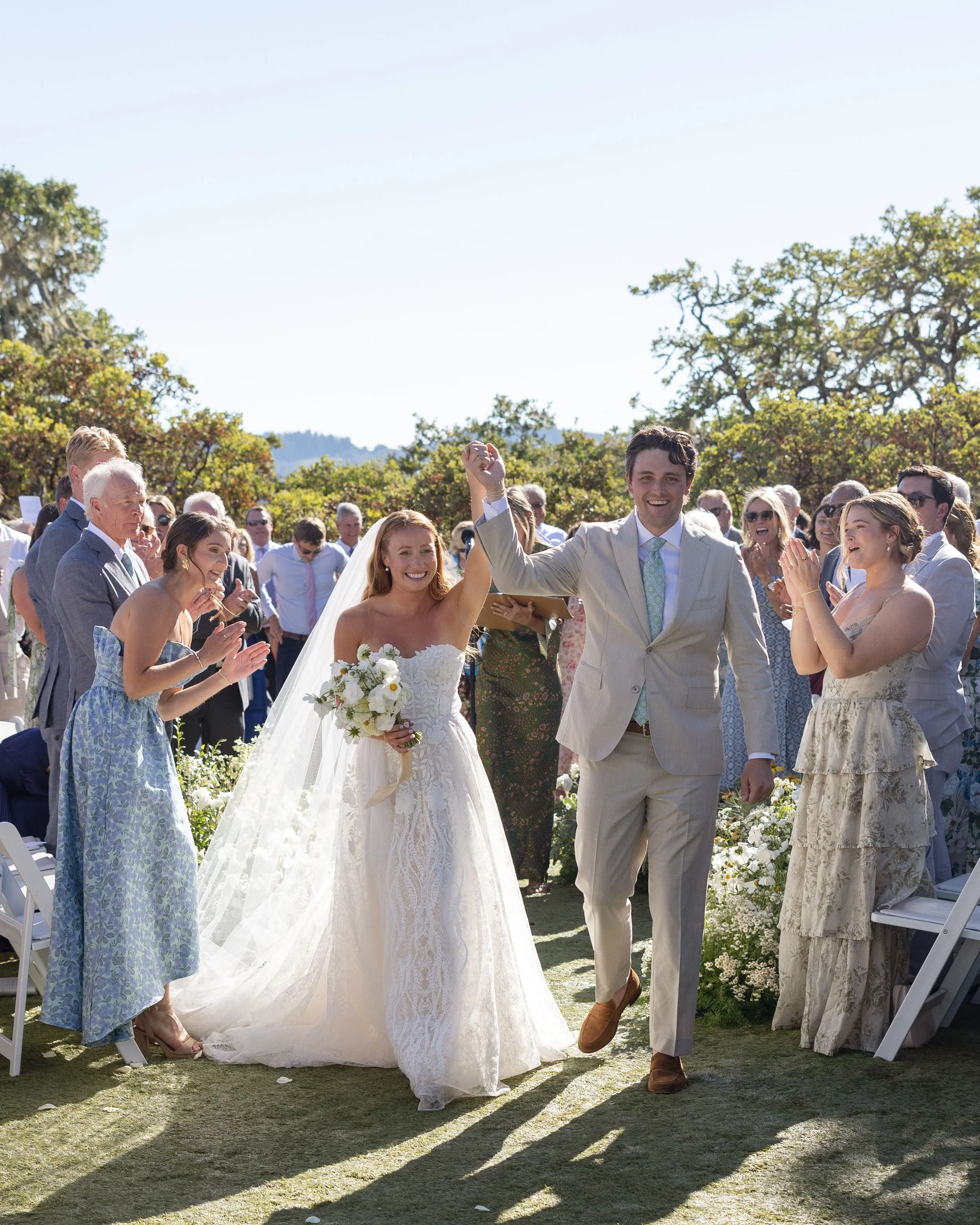 A wedding ceremony outdoors with a bride and groom walking down the aisle, surrounded by guests. The bride wears a white lace wedding dress and holds a bouquet, while the groom wears a light beige suit with a pastel tie. The guests celebrate as they 