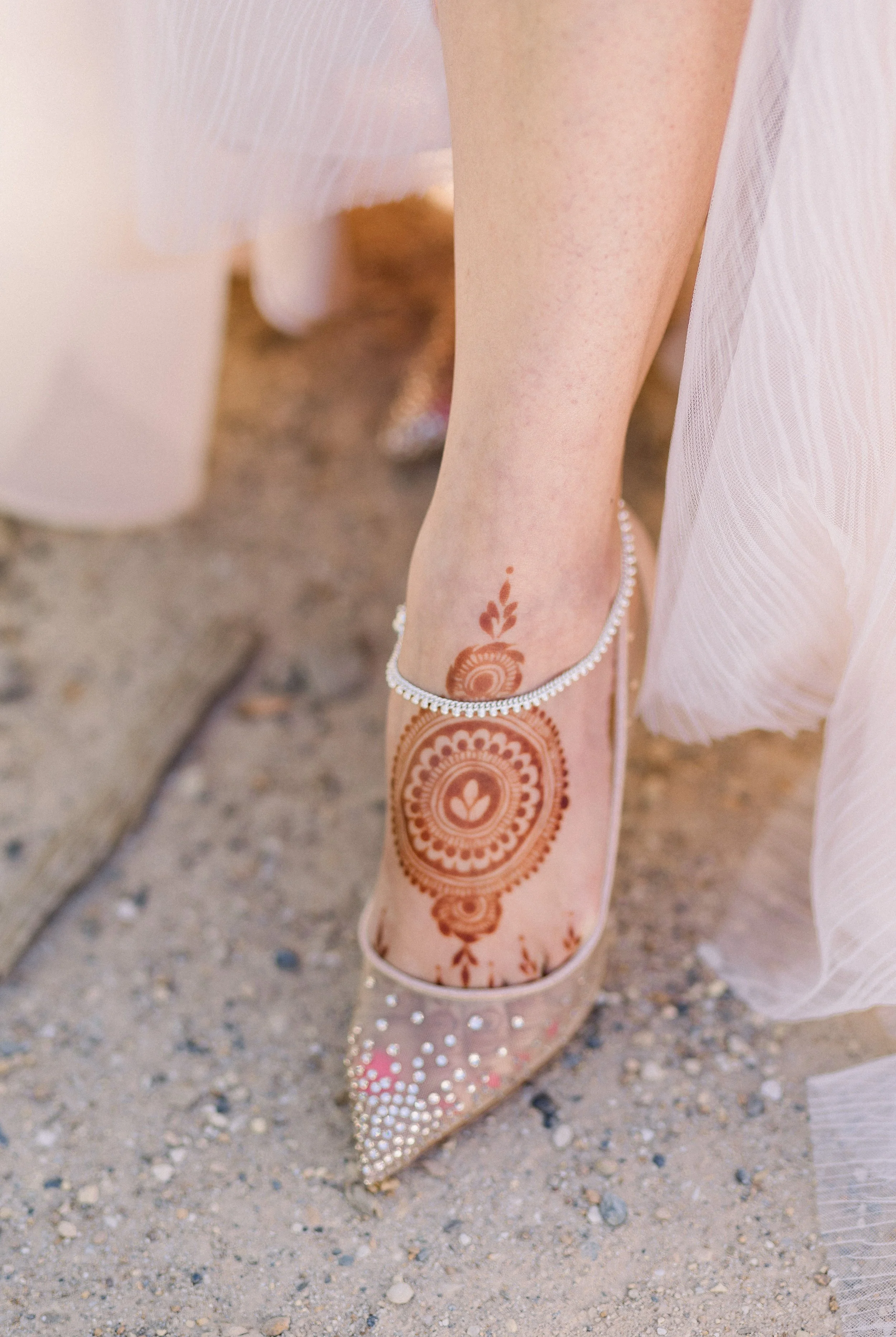Close-up of a person wearing a party dress with henna tattoo on their leg, decorated with rhinestones, and white high heels with rhinestone embellishments.