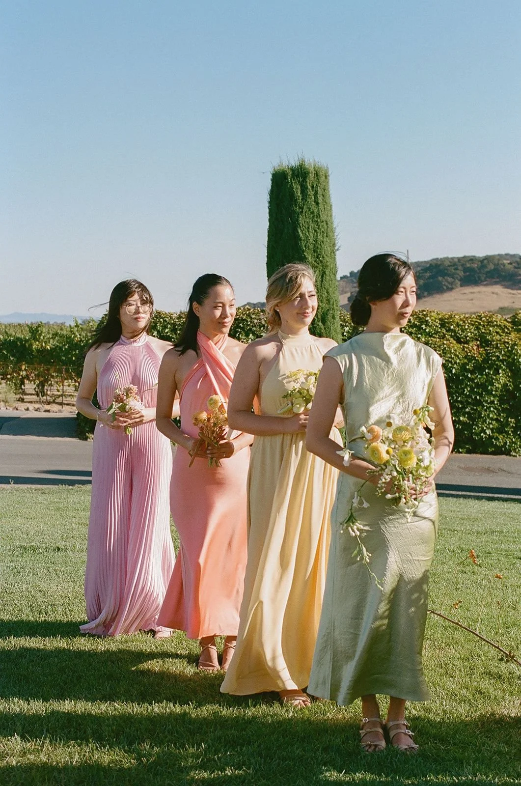 Four women standing in a line outdoors, each holding a bouquet of flowers, wearing pastel-colored dresses, with a tall green shrub and vineyard in the background on a sunny day.