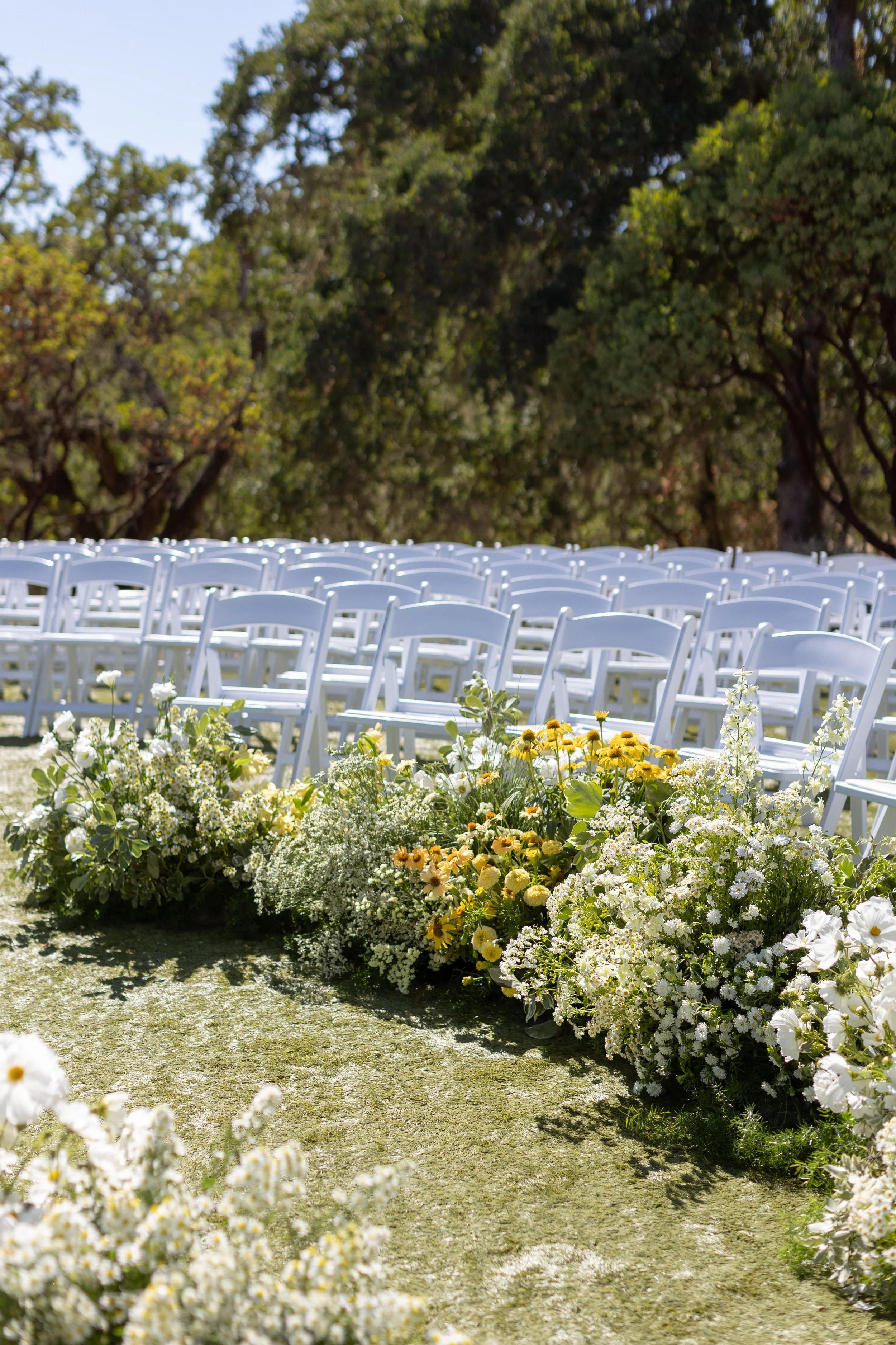 Empty white chairs arranged outdoors for a wedding ceremony with a floral aisle in the foreground and trees in the background.