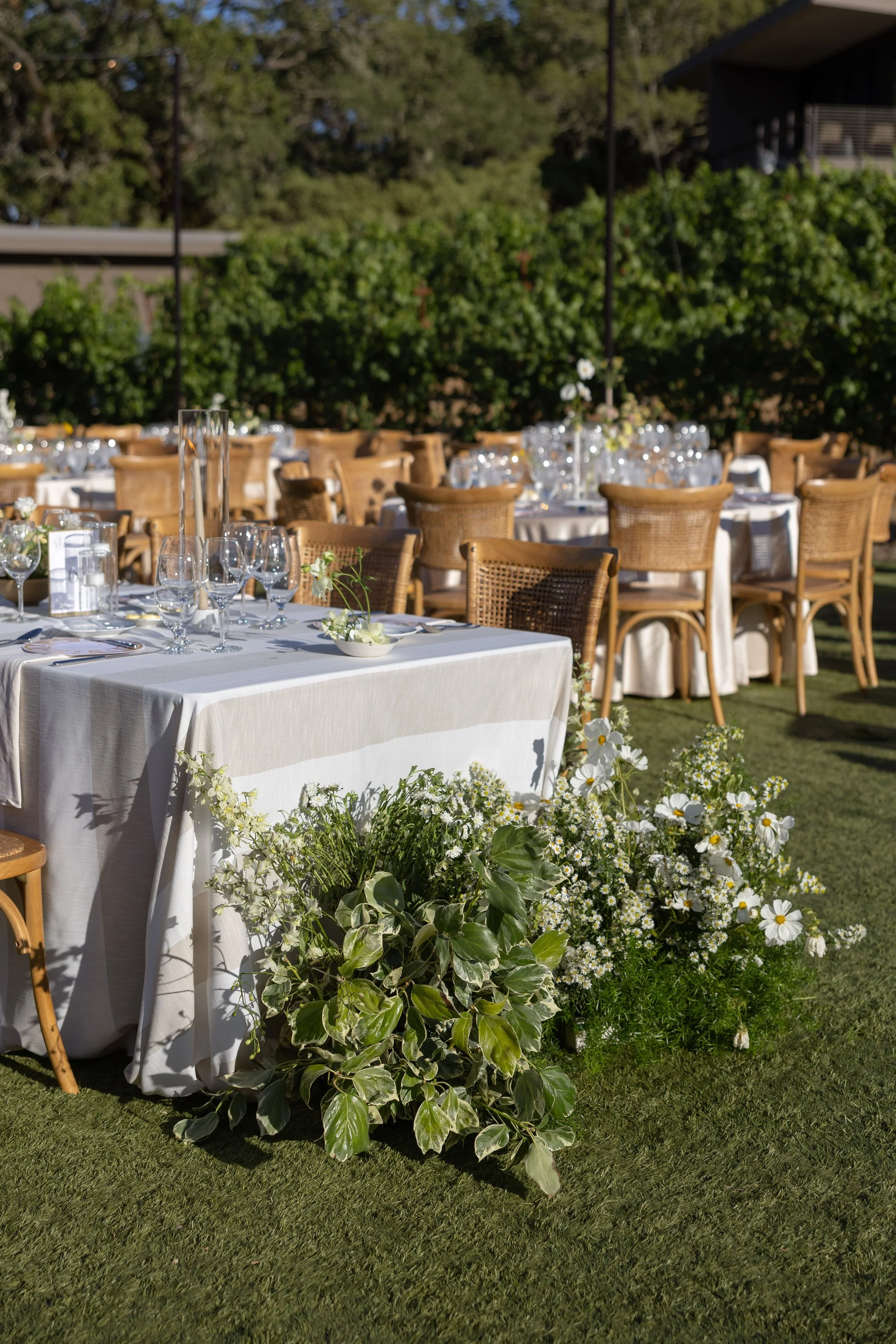 Outdoor event setup with a long table covered in white tablecloth, surrounded by wooden chairs, and decorated with glassware and floral arrangements, with greenery and trees in the background.