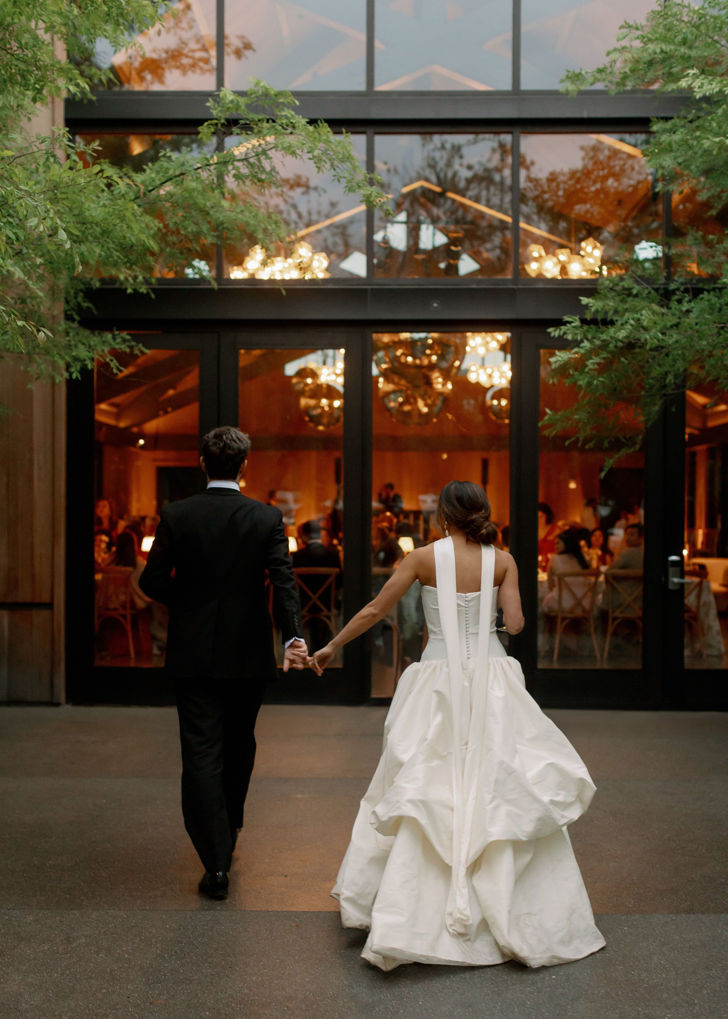A bride and groom walking hand in hand towards a wedding reception venue, seen from behind. The bride is in a white gown, and the groom is in a black suit. The venue has glass doors, and warm lights are visible inside, with chandeliers hanging from t