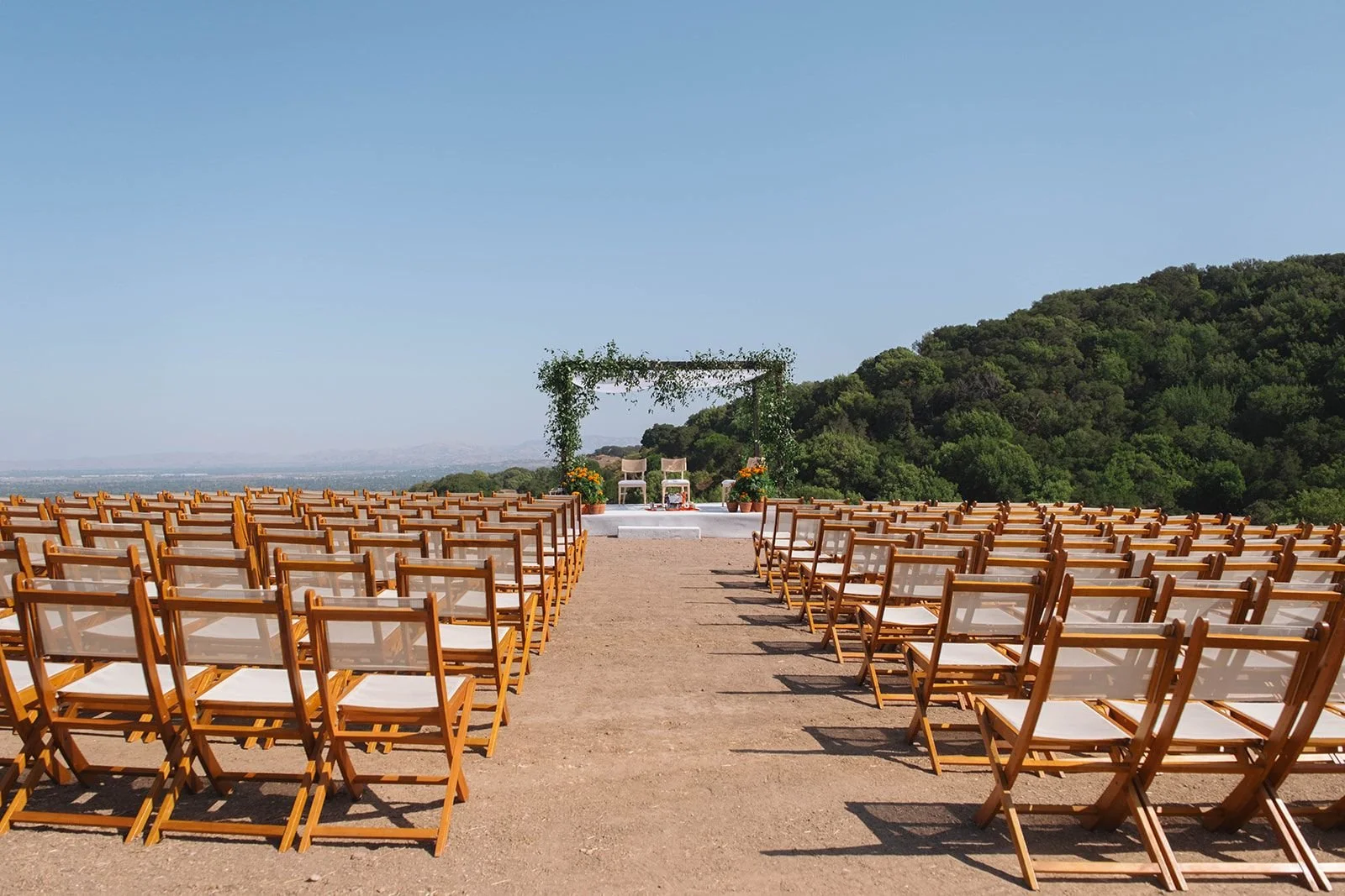 Outdoor wedding setup with rows of wooden chairs facing a decorated arch with green foliage and flowers, on a dirt ground with hills and trees in the background under a clear blue sky.