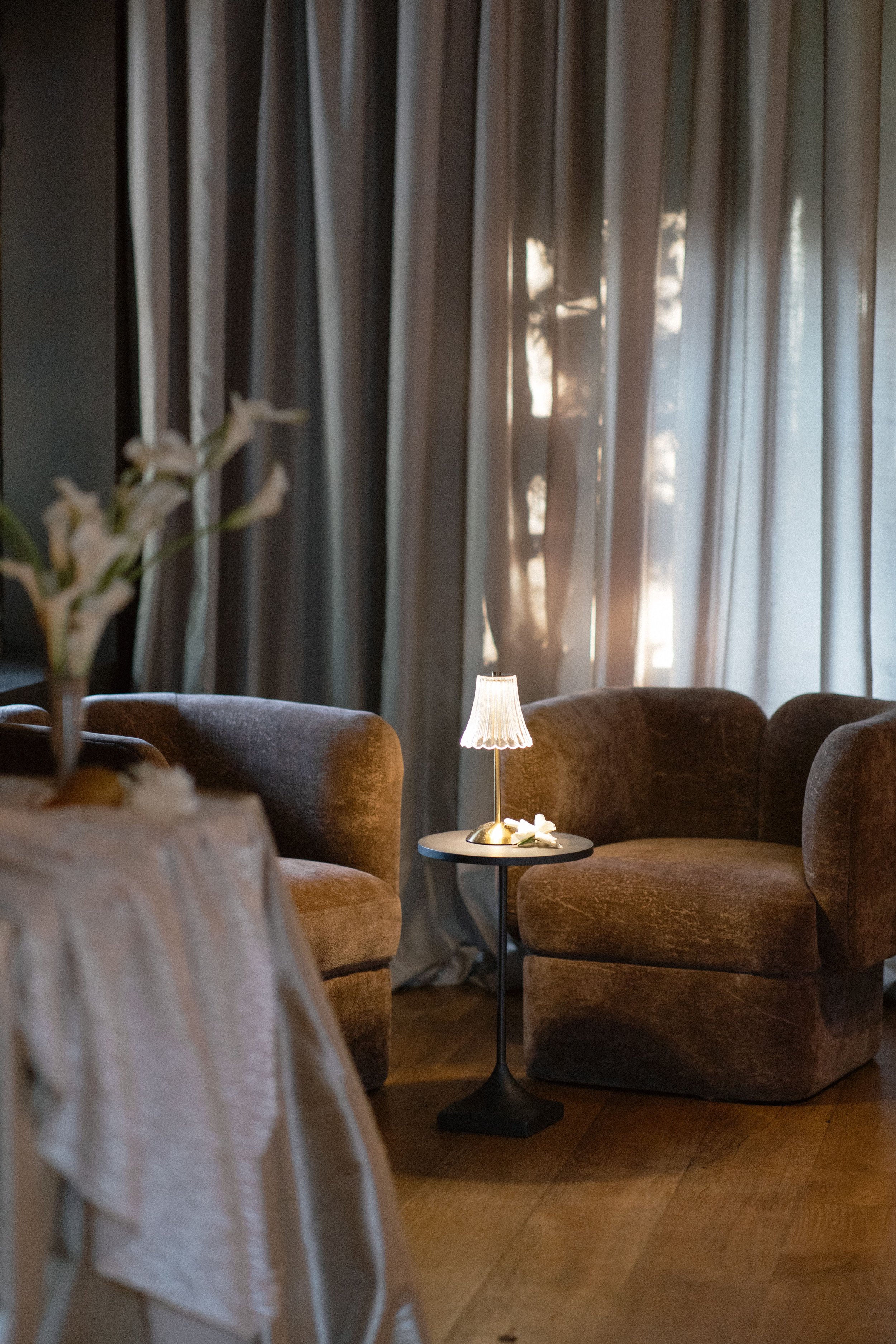 A cozy living room with two brown upholstered armchairs arranged around a small black side table. A small lamp with a pleated shade sits on the table, casting a warm glow. Behind, there are long, beige curtains with light filtering through them, crea