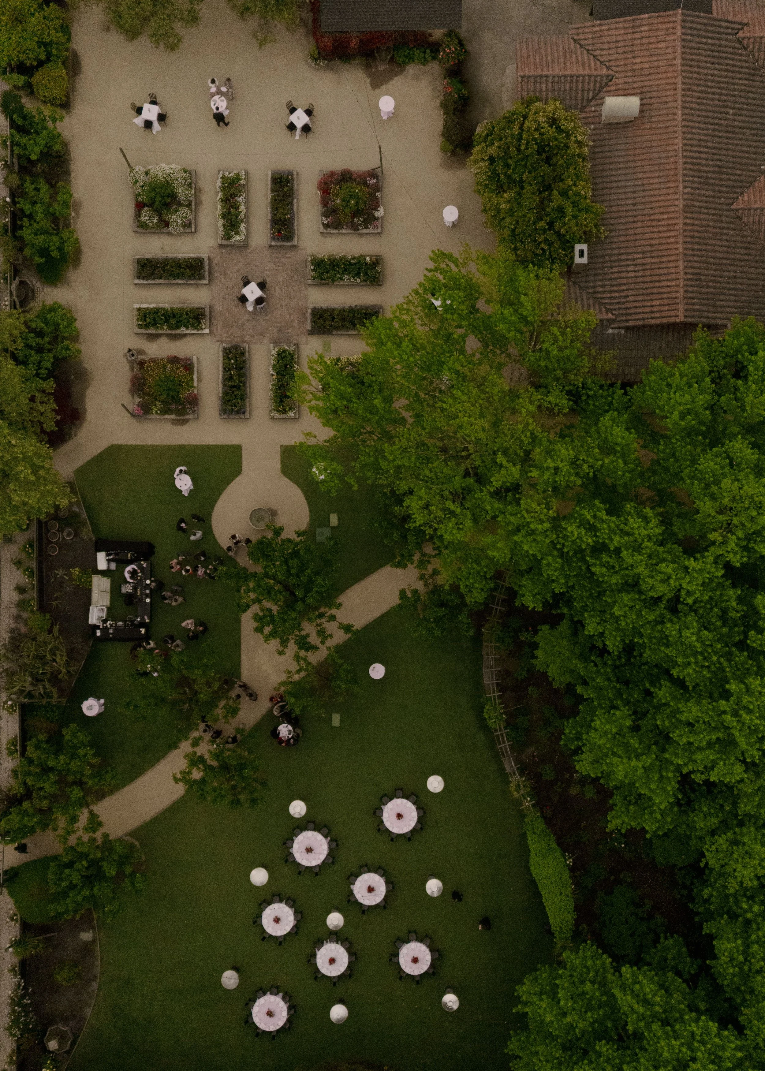 An aerial view of an outdoor event area with tables covered in white tablecloths, surrounded by chairs. There are garden beds with flowers and a grassy lawn with several large umbrellas and groups of people. A building with a tiled roof is nearby, an