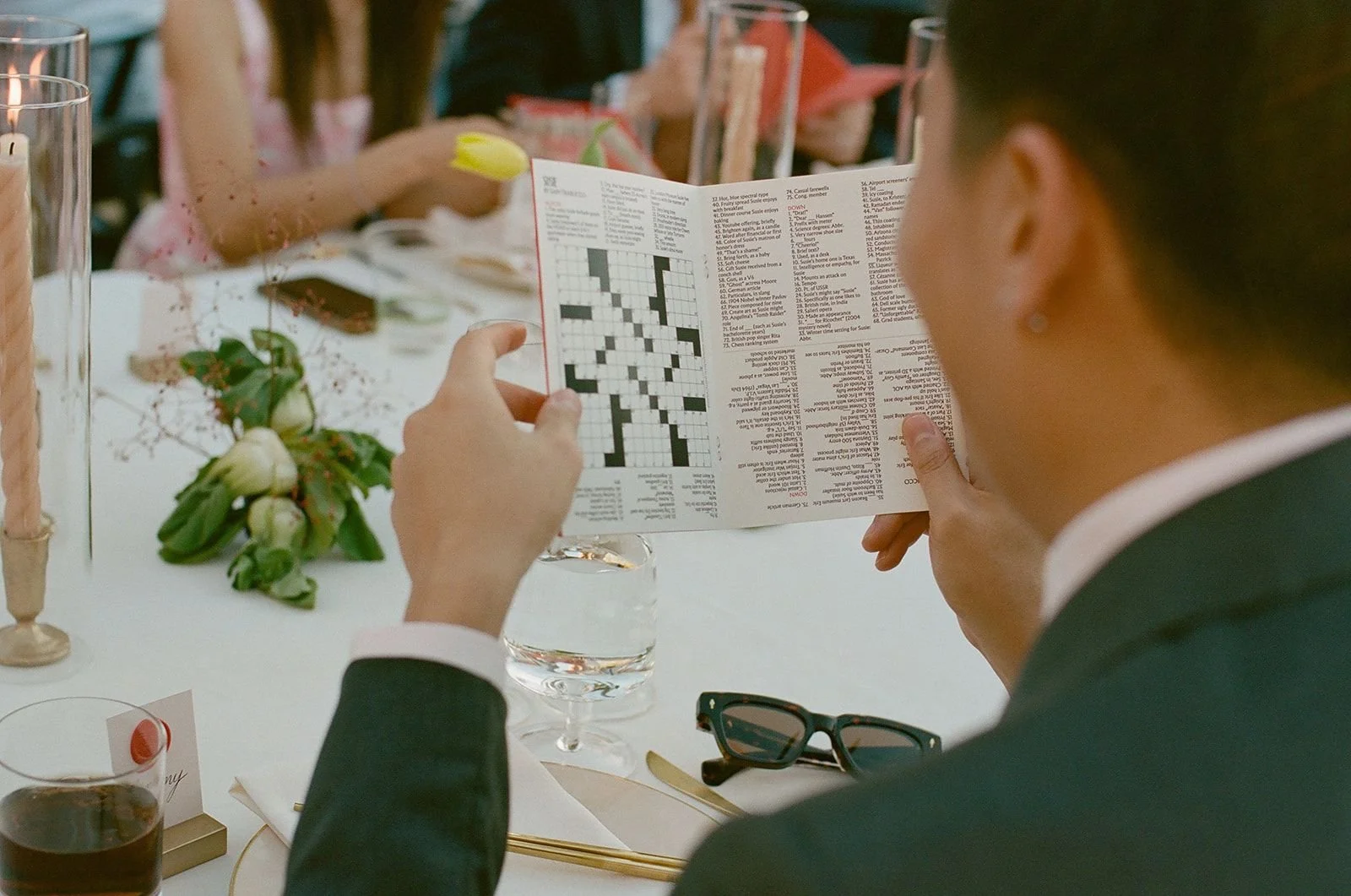 A person in a suit is sitting at a table, holding a crossword puzzle or newspaper, with glasses and a glass of water on the table. There is a floral centerpiece and other people in the background, indicating a social or formal event.
