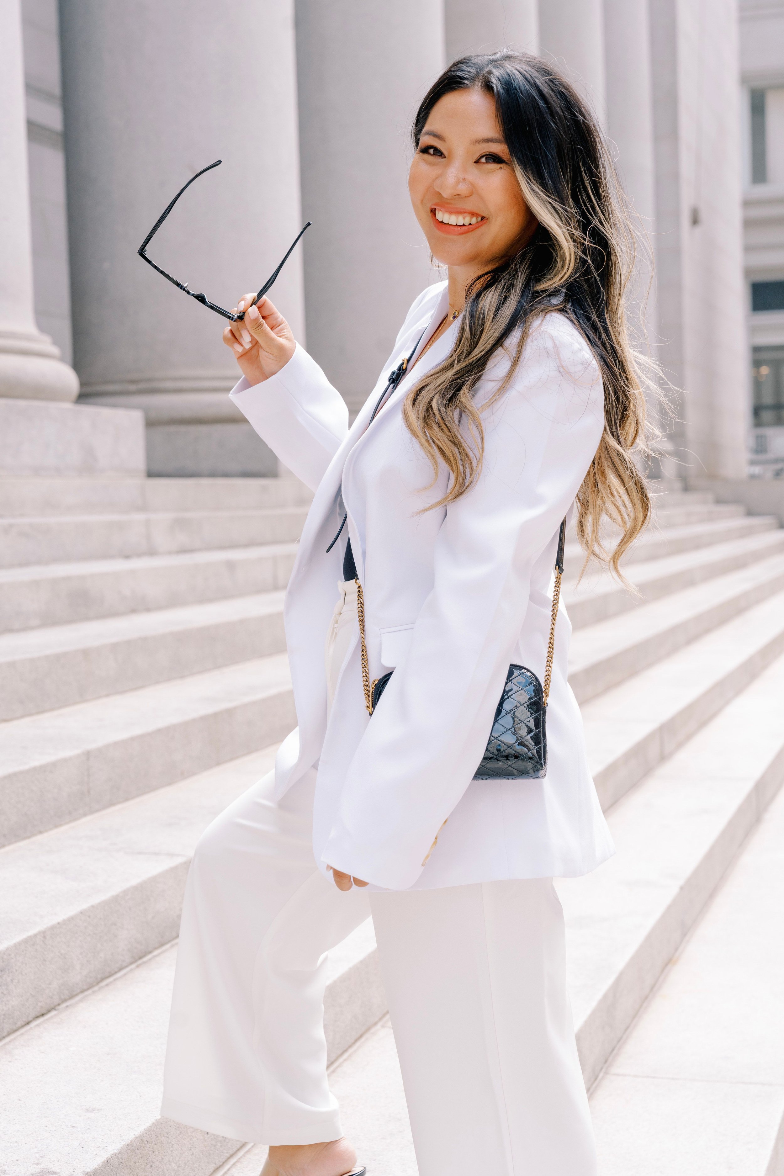 A woman with long, wavy hair, wearing a white suit, smiling, holding sunglasses in her right hand, standing on the steps of a large, classical-style building with columns.