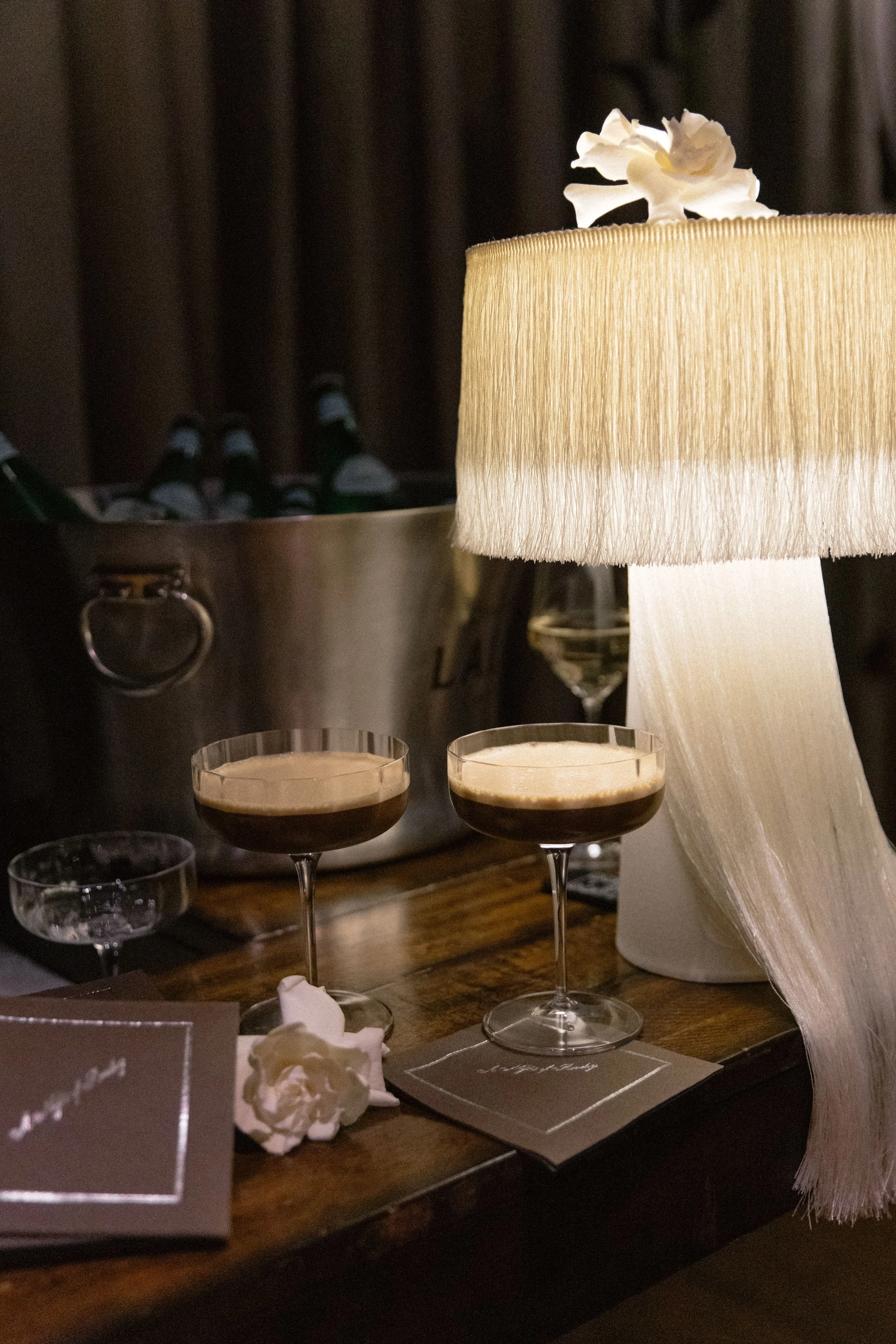 Two cocktails in coupe glasses in front of a cream-colored lampshade with a white flower on top. There is a metal bucket with bottles in the background, and a wooden table with some papers and a card on it.
