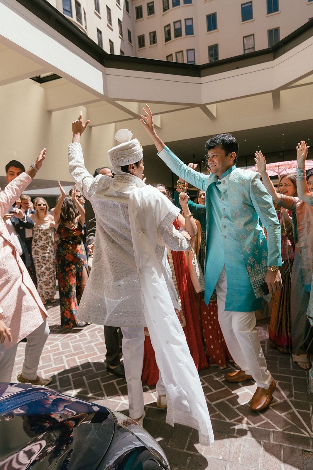 People celebrating at a wedding or cultural event, dancing and high-fiving outdoors under a modern building with multiple windows.