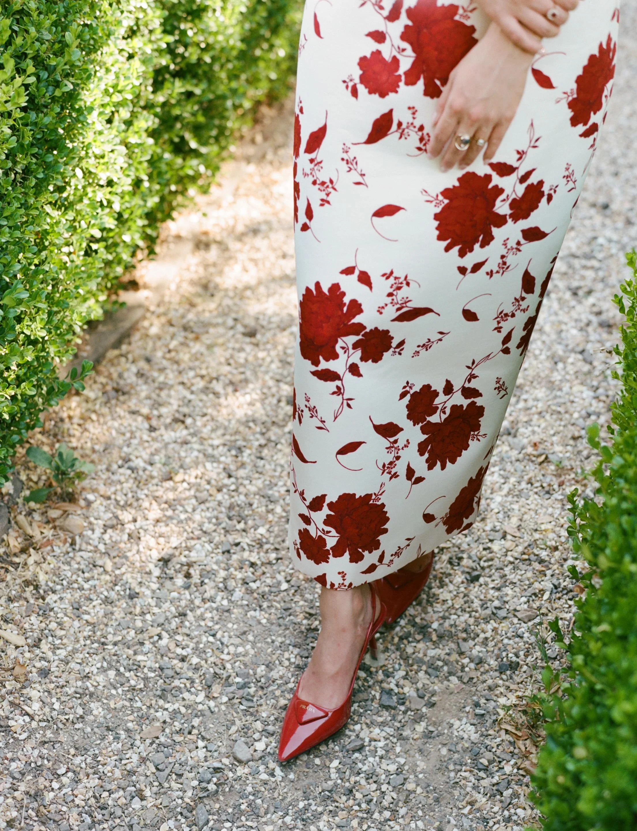 A woman wearing a white dress with red floral pattern, red high-heeled shoes, and rings on her fingers, standing on a gravel path between green bushes.