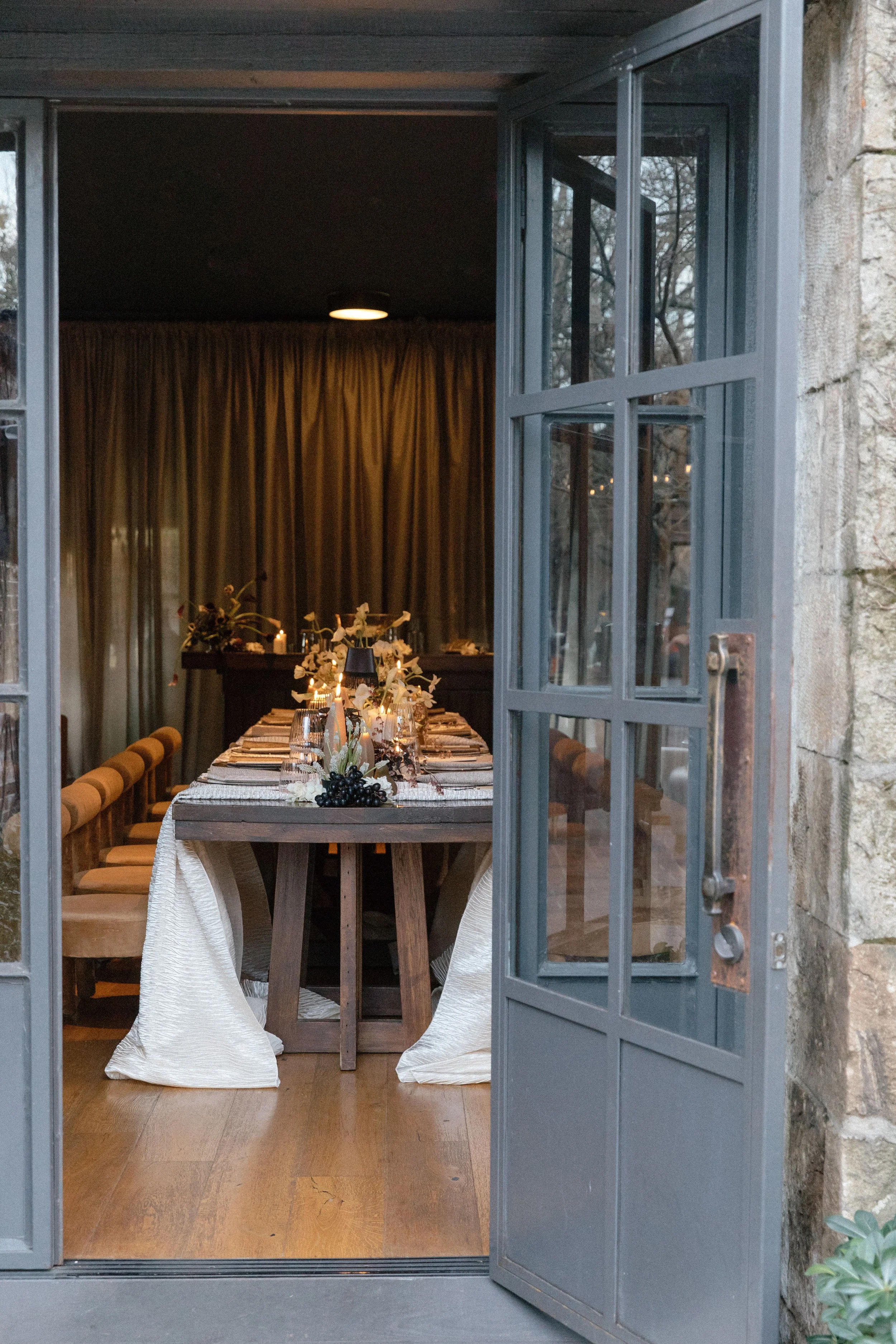 Open door revealing a dining room with a long table decorated with flowers, candles, and wine glasses. The room has wooden floors, beige curtain, and soft lighting.