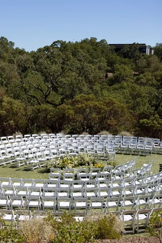 Outdoor wedding ceremony setup with white chairs arranged in an arc, surrounded by greenery and trees on a hillside.