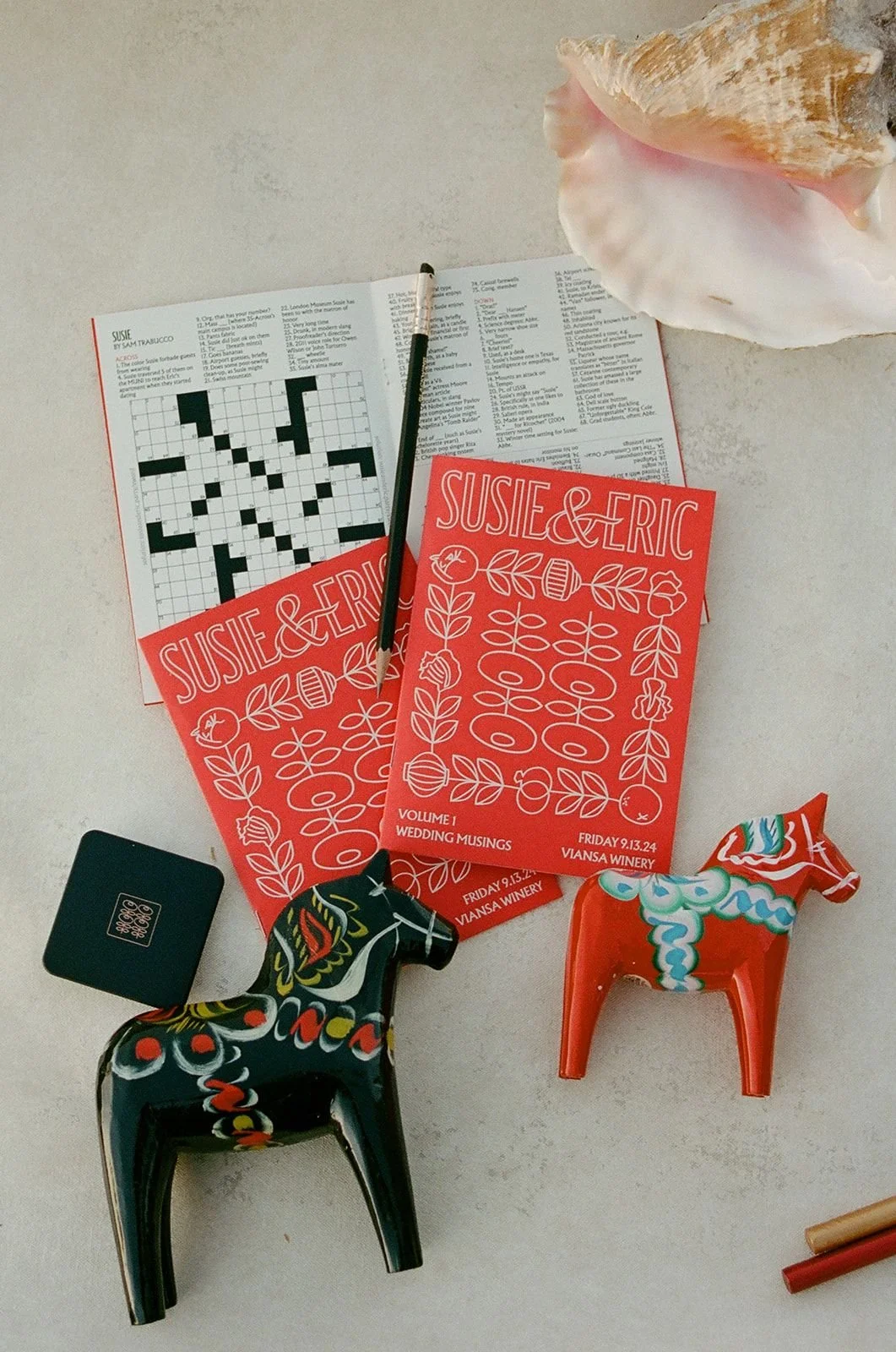 Colorful paperboard horse figurines, a printed crossword puzzle, event flyers for Susie & Eric wedding musings, a small black device, and a conch shell on a light-colored surface.
