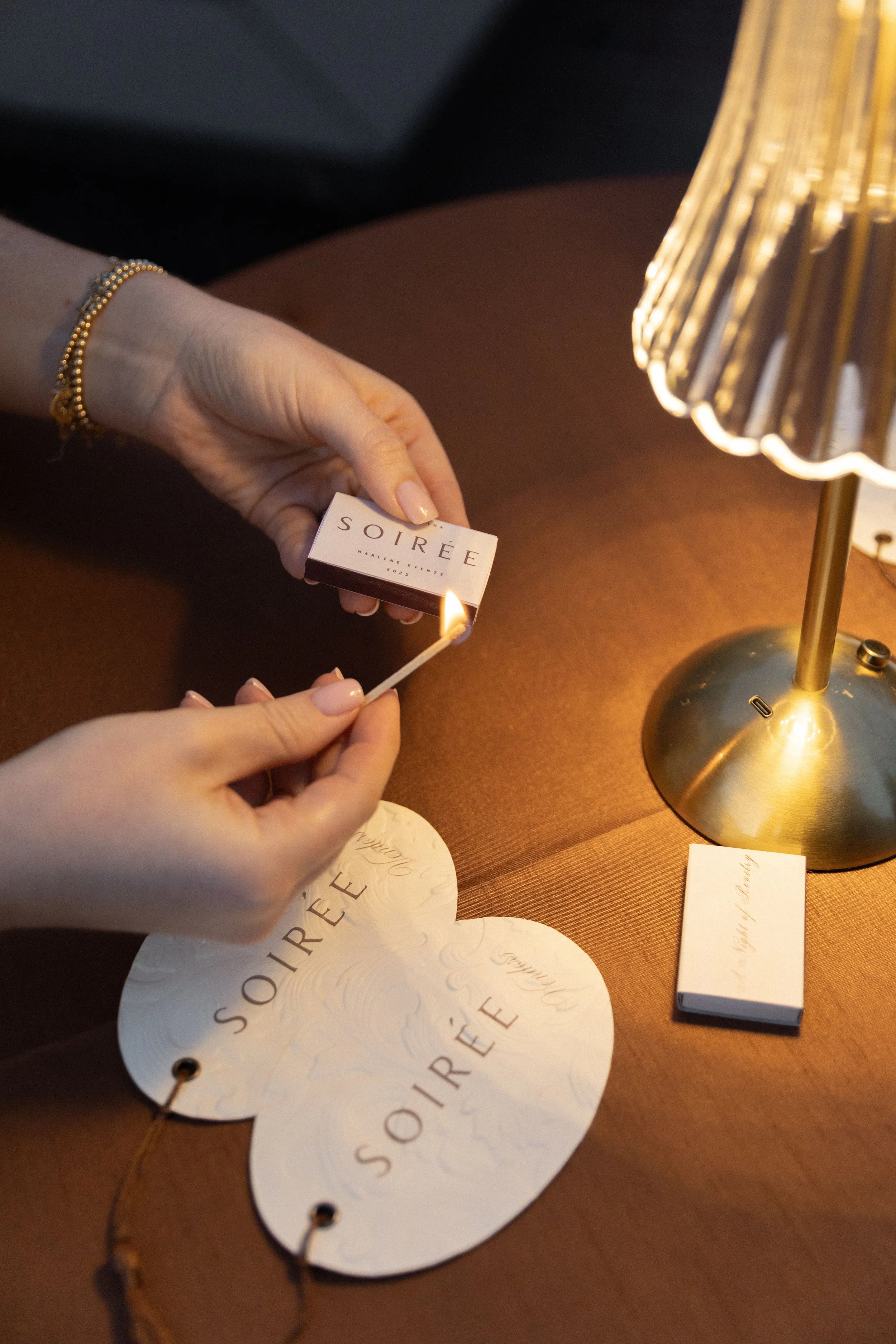Person holding a matchbox labeled 'SOIRÉE' while lighting a match near a gold table lamp, with branded paper items on a brown table.