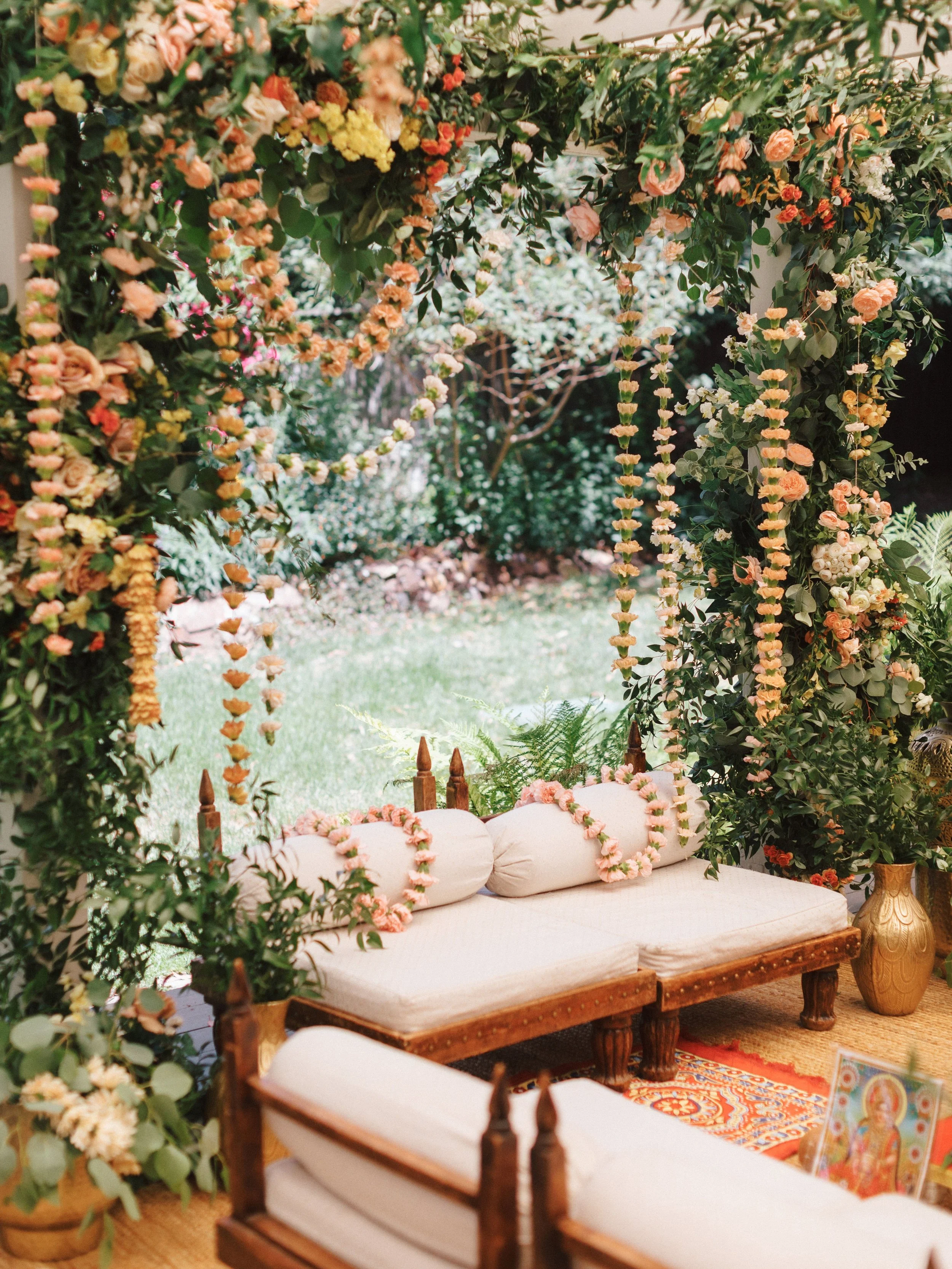 An outdoor wedding setup with a cream cushioned wooden bench decorated with pink floral garland, surrounded by lush green foliage and flowering plants, set on a decorative rug.