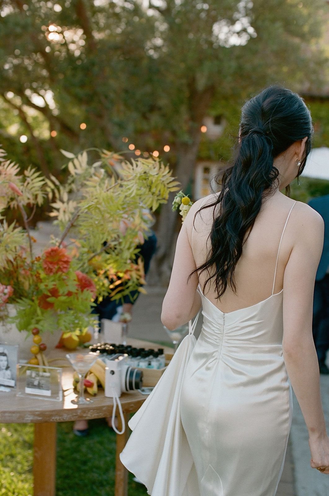 A woman with long black wavy hair wearing a cream-colored backless dress at an outdoor event, with a table of decorative items and greenery in the background.