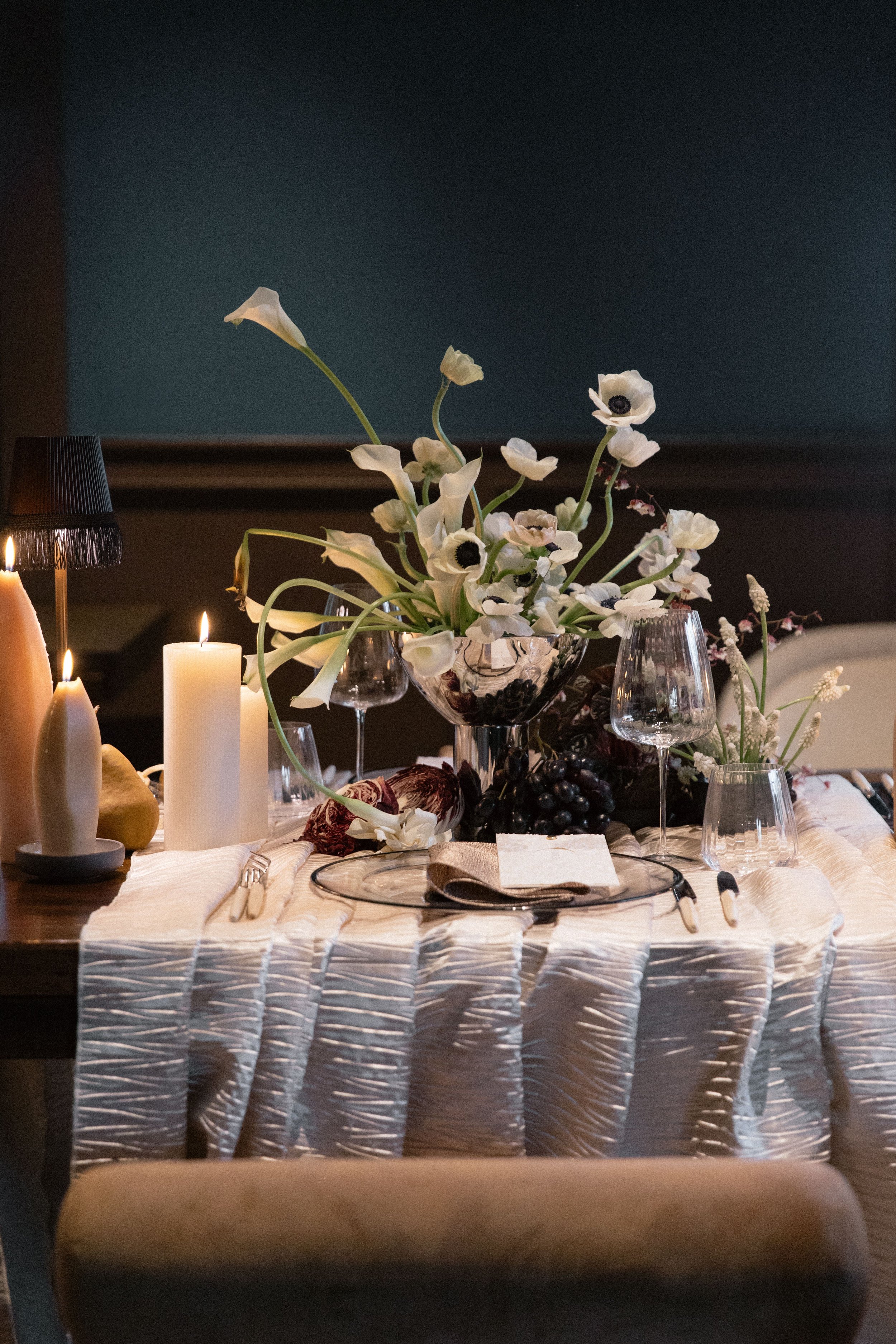 A table set for a formal dinner with a cream-colored tablecloth, candles, glassware, and a floral centerpiece featuring white anemones and other flowers.