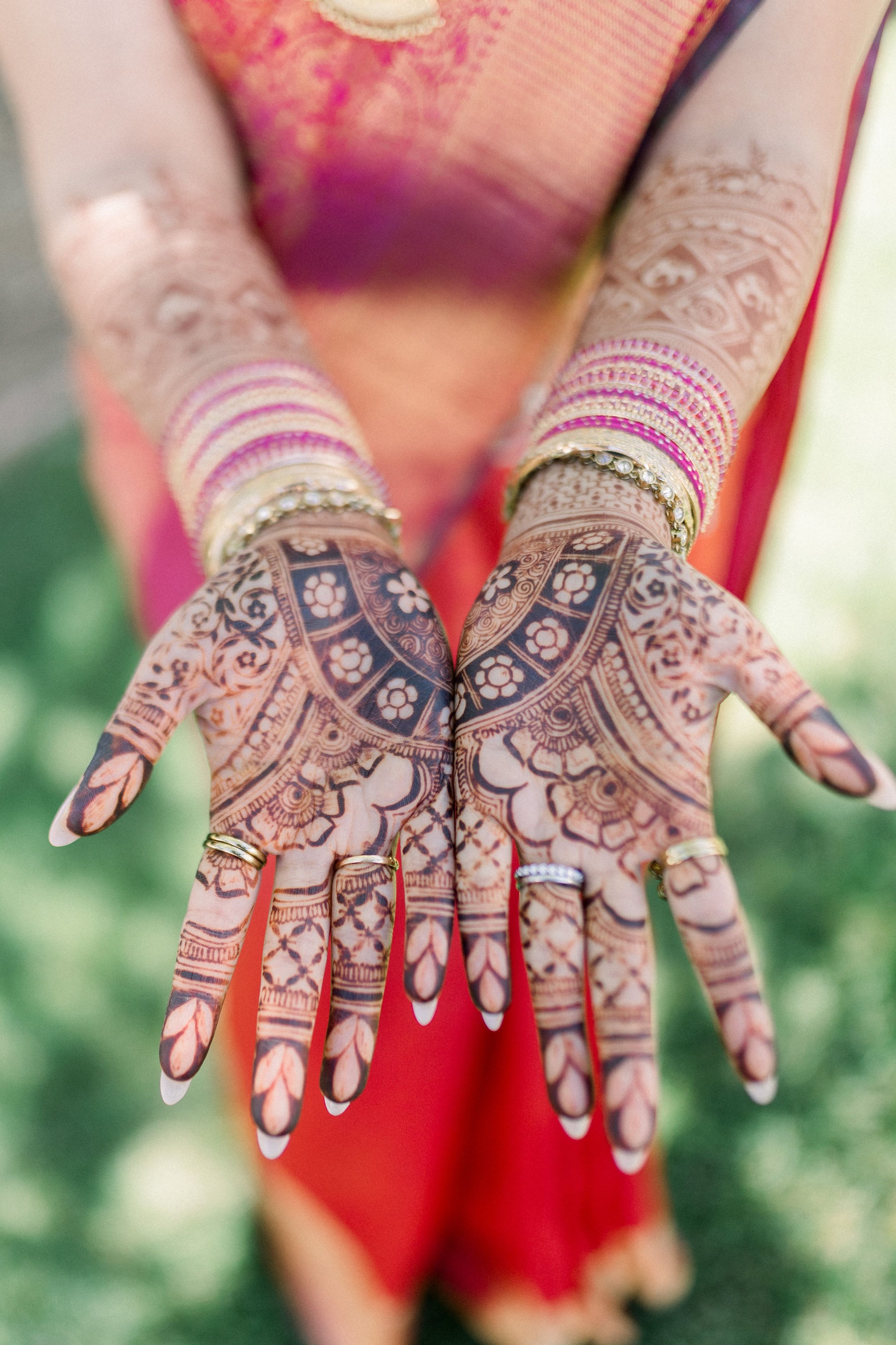 Close-up of a person's hands with intricate henna designs, wearing multiple colorful bangles and rings, with a blurred background.