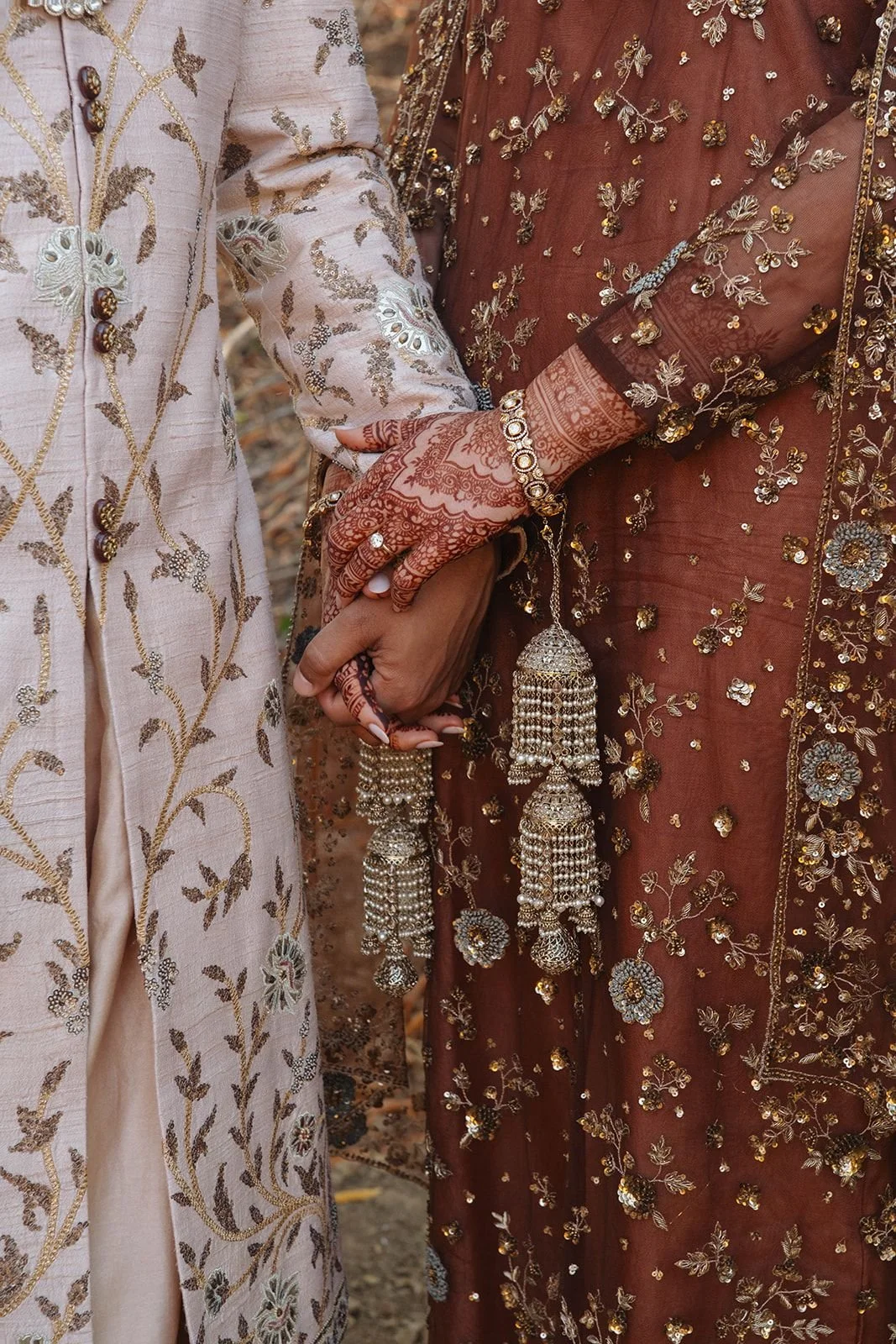 Close-up of two people holding hands, dressed in ornate traditional Indian clothing with intricate embroidery and jewelry, including bangles and embellished sleeves.