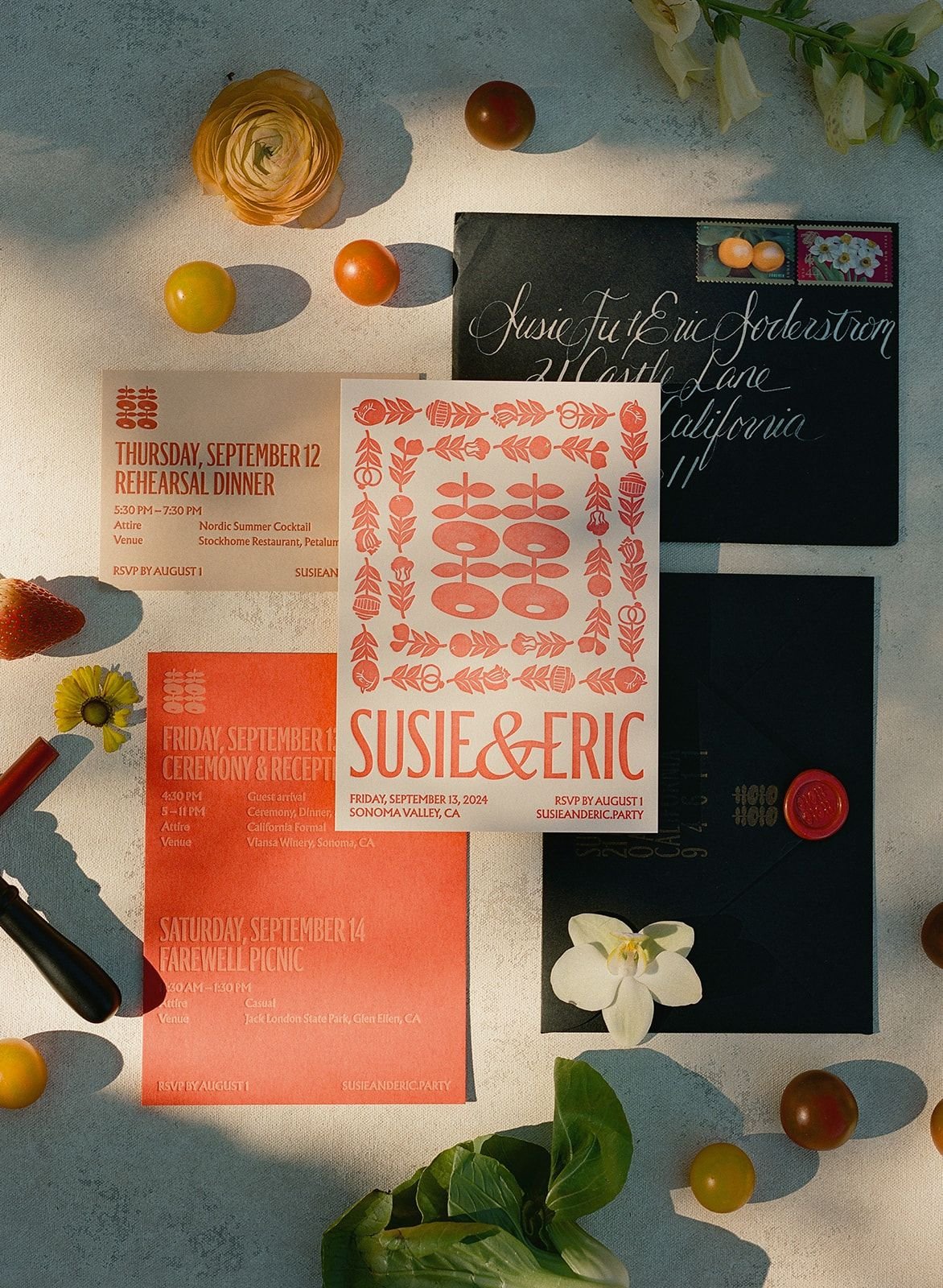 Wedding invitation suite with bright orange and pink hues, featuring the names Susie and Eric and details of their wedding events from September 12 to 14, 2024, with floral and decorative elements. The suite is surrounded by small cherry tomatoes, fl