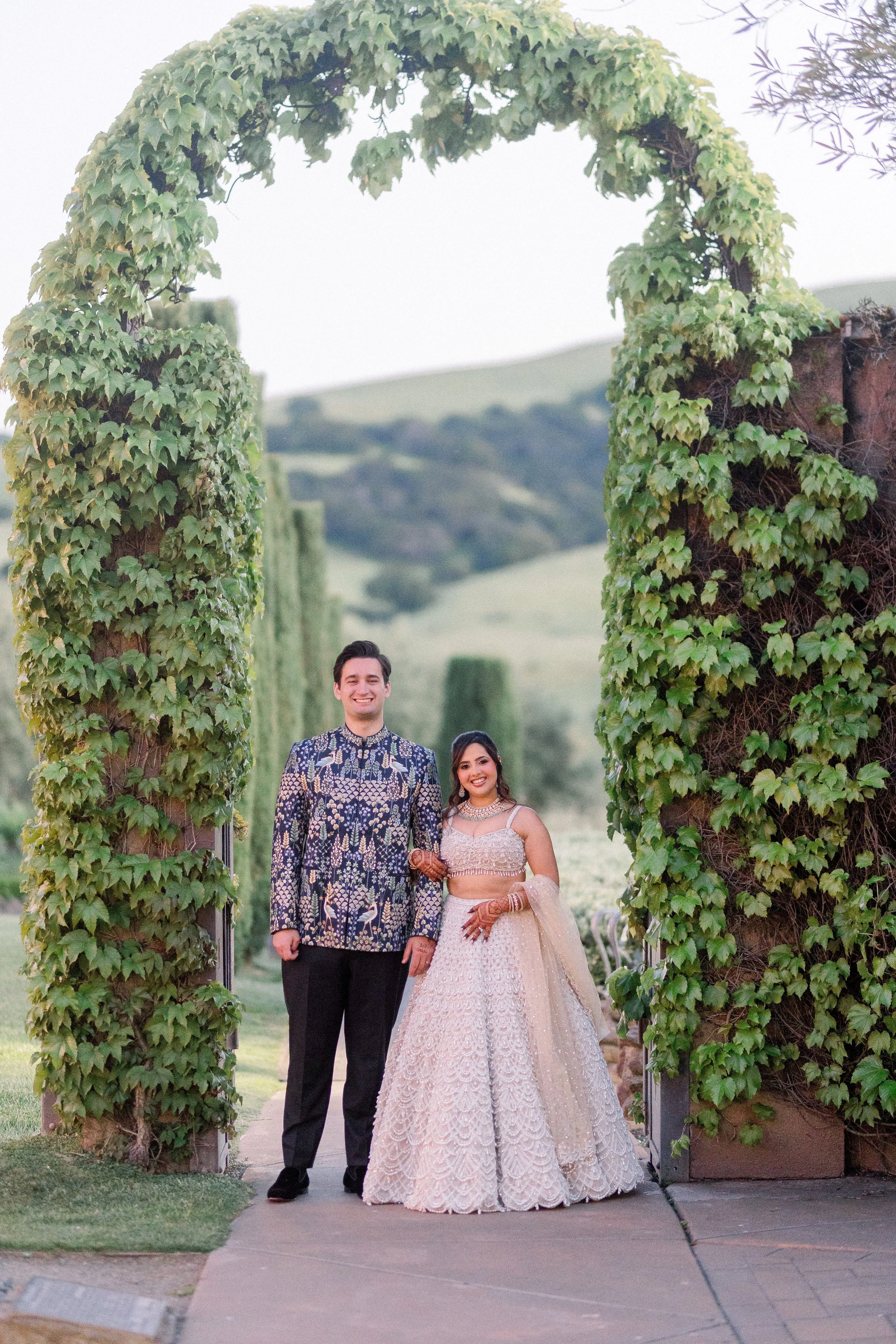 A smiling man and woman dressed in traditional Indian attire standing under a large leafy arch at sunset, with green hills in the background.