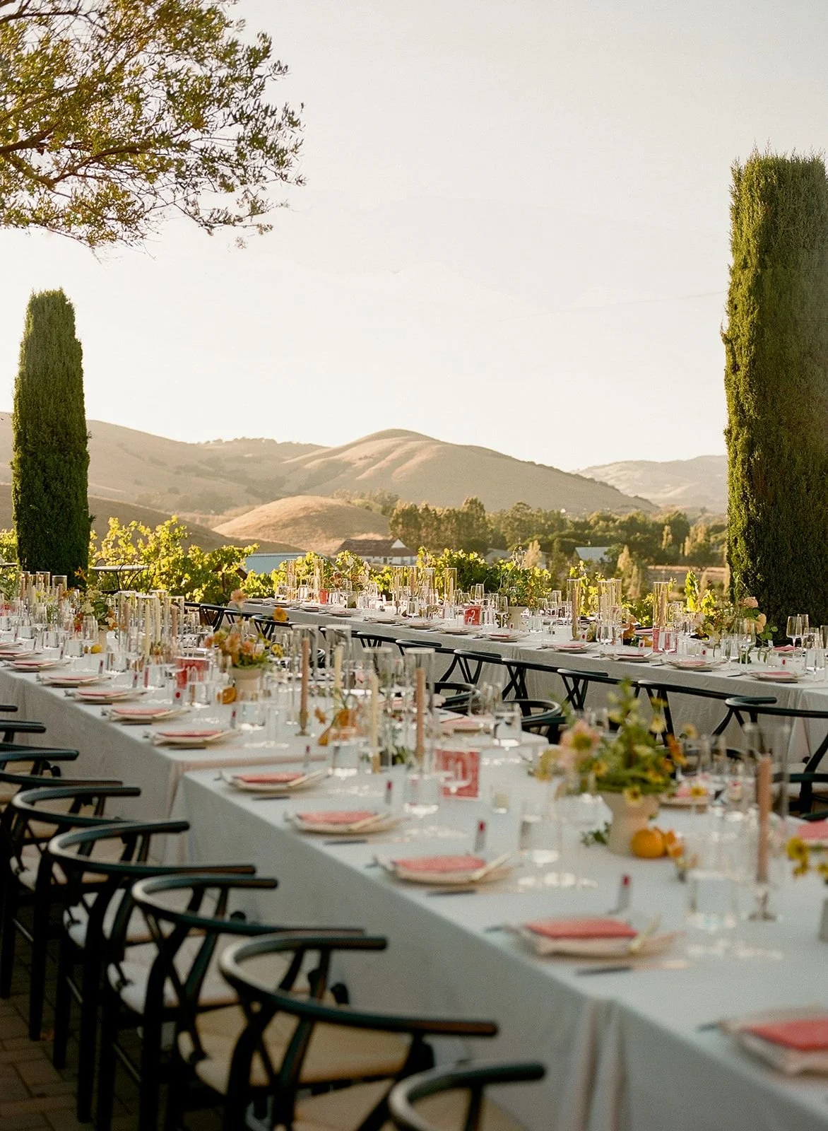 Long banquet table set up outdoors with elegant tableware, candles, and floral arrangements, overlooking hills and trees in the background during sunset.