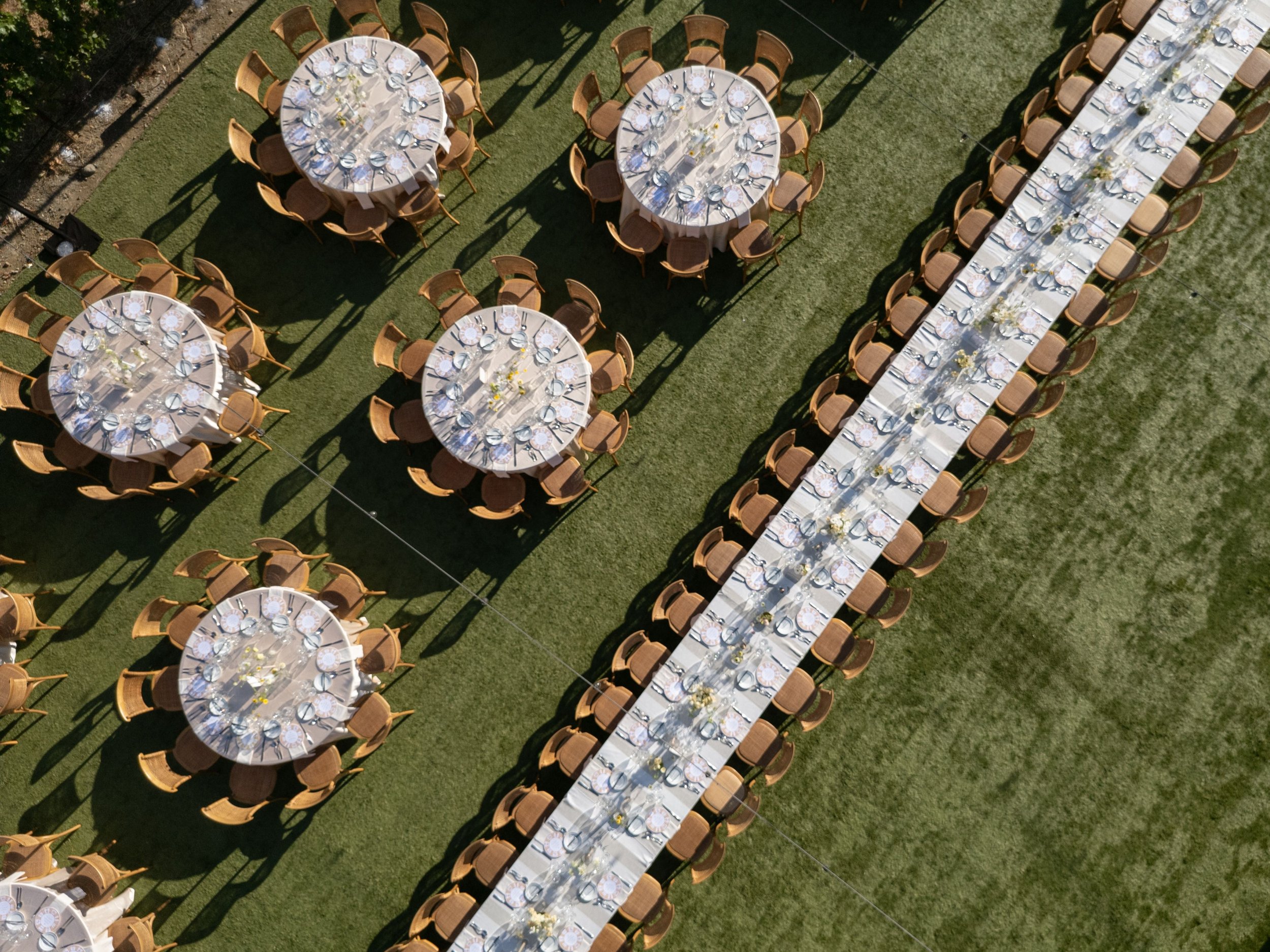 An outdoor event setup with round and long banquet tables decorated for a celebration or wedding, featuring place settings, glassware, and floral centerpieces, all on a grassy area under bright sunlight.