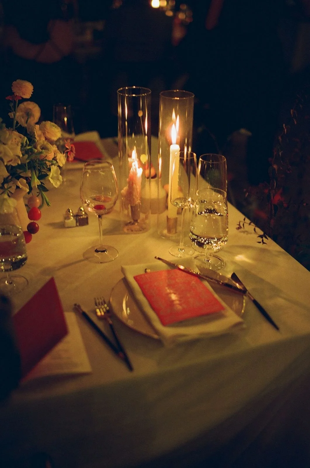 Elegant dinner table set with candles, wine glasses, a floral centerpiece, and folded napkins in a dimly lit setting.