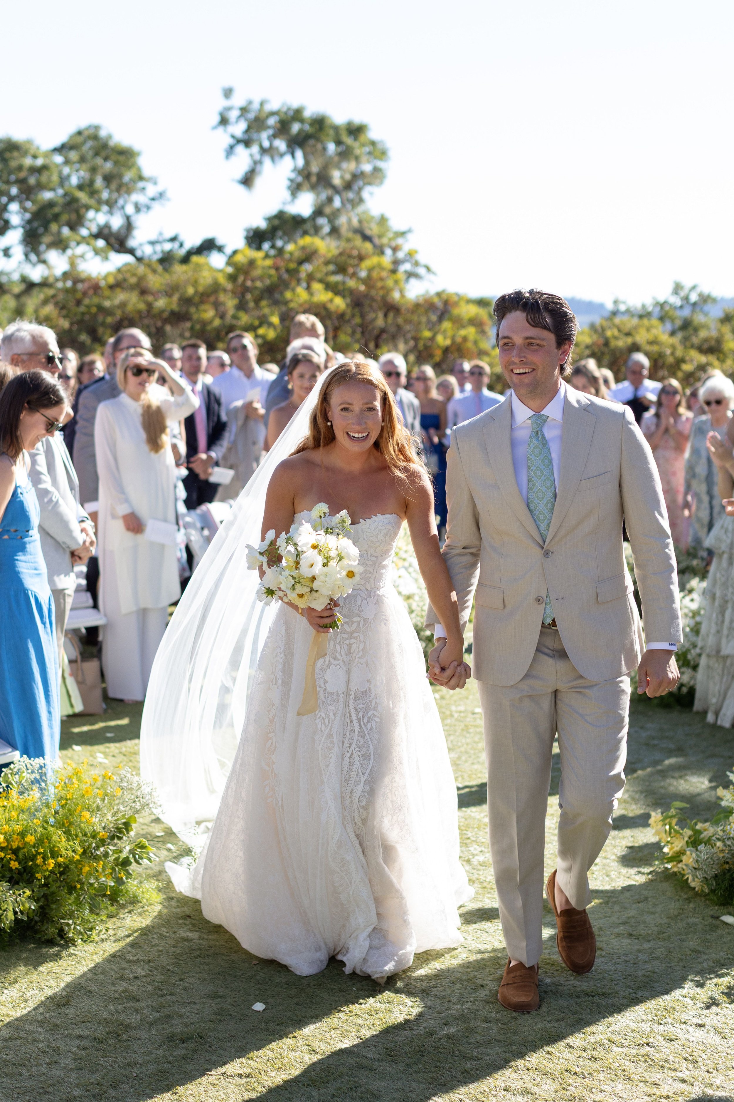 A bride and groom walk hand in hand outdoors after their wedding ceremony, smiling and holding a bouquet of white flowers, with guests in the background celebrating.