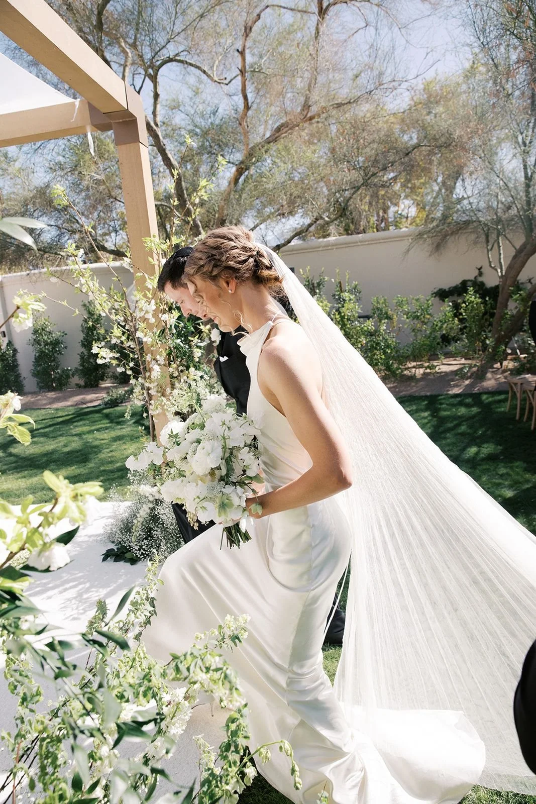 Bride in white wedding dress holding bouquet, smiling, outdoor wedding ceremony, green grass, trees, sunny day.