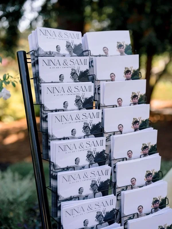 Display stand with wedding programs or invitations labeled 'Nina & Sami' arranged in rows outdoors.
