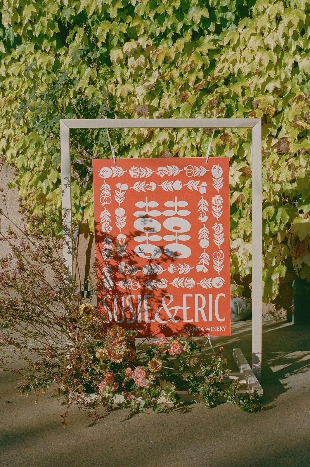 A sign with white floral patterns and the text "SUSIE & ERIC" hangs from a white frame. The background is a wall covered with green ivy leaves, and there are pink flowers at the base of the sign.