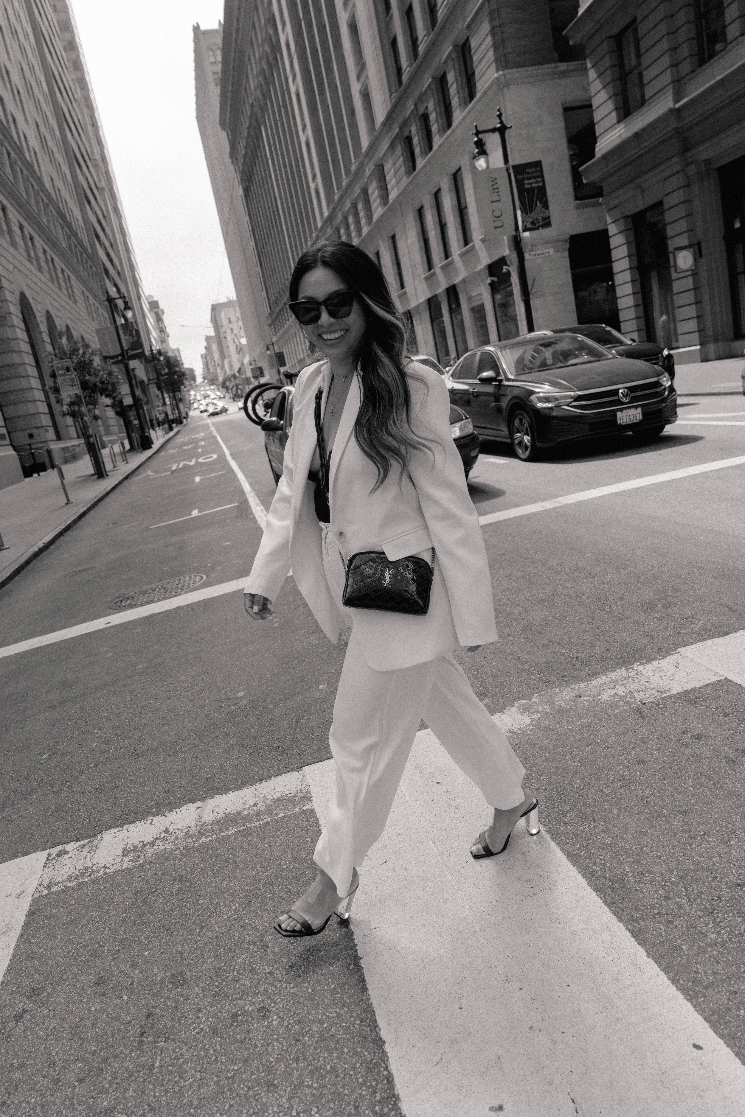 A woman wearing a light-colored blazer, white pants, and high-heeled sandals walking in an urban street, smiling and wearing sunglasses. Black and white photo.