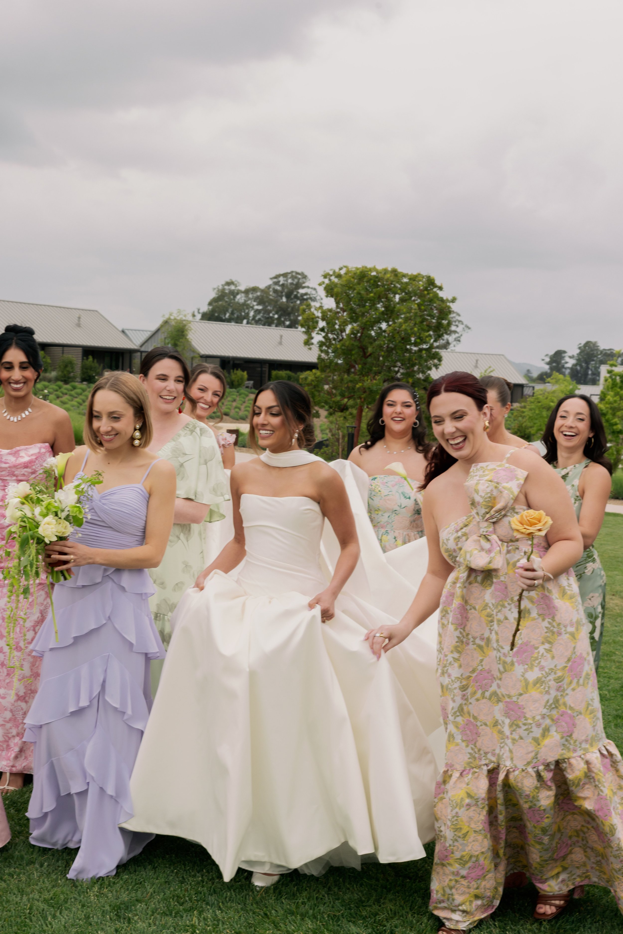 A group of women in colorful dresses are standing outdoors on a grassy area, smiling, holding hands, and laughing. The background shows houses, trees, and an overcast sky.