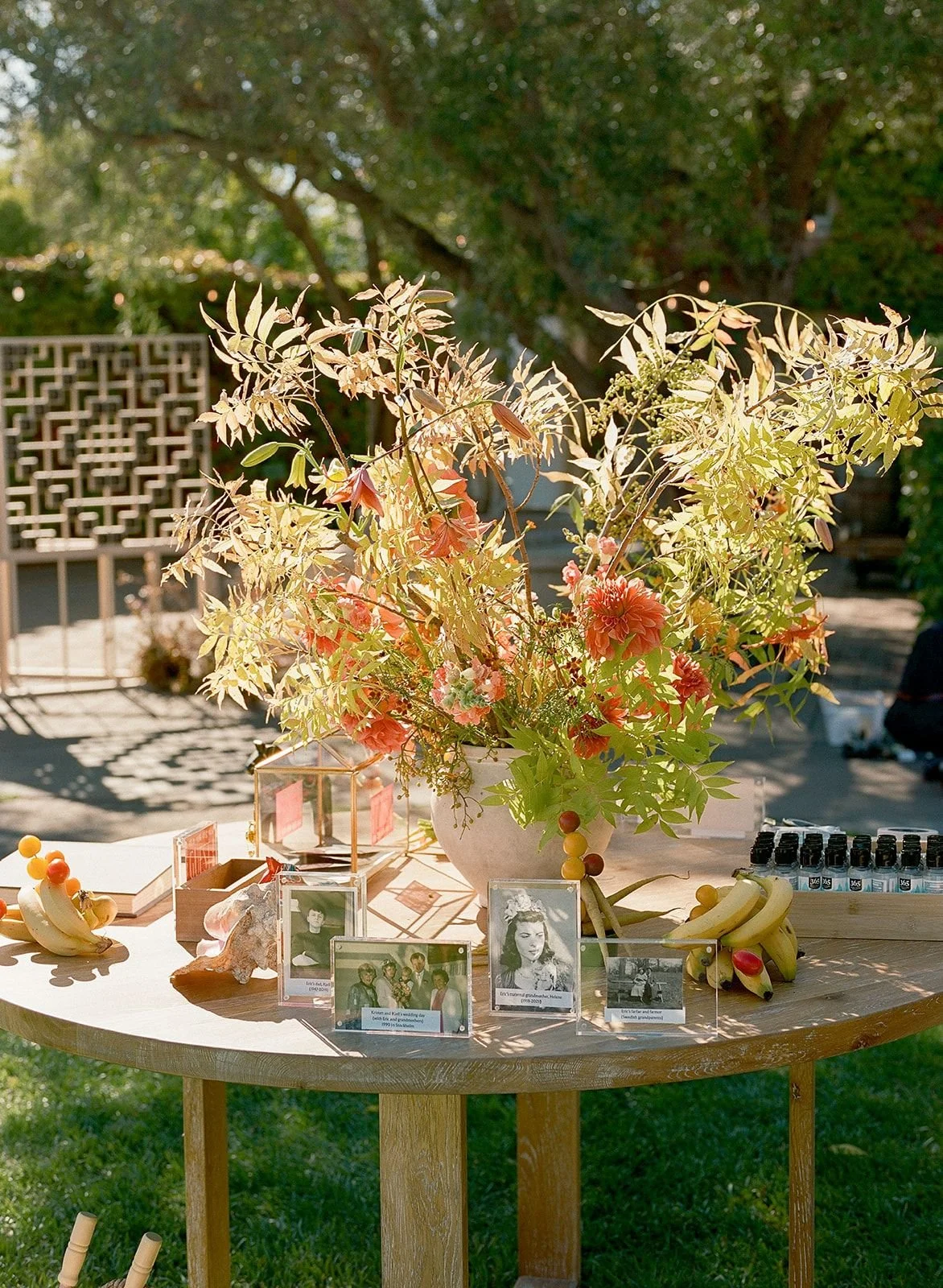 A round wooden table with a large floral arrangement in a white vase, surrounded by framed photos, bananas, and small decorative items, set outdoors in a garden.