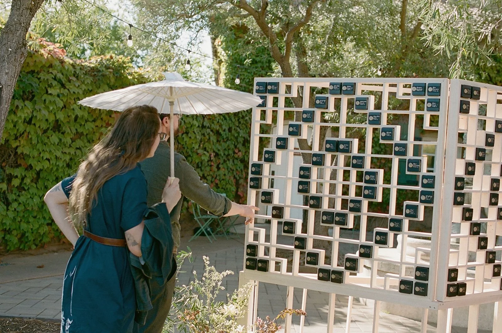 People playing a large grid-based game outdoors, with one person holding an umbrella, surrounded by trees and string lights.