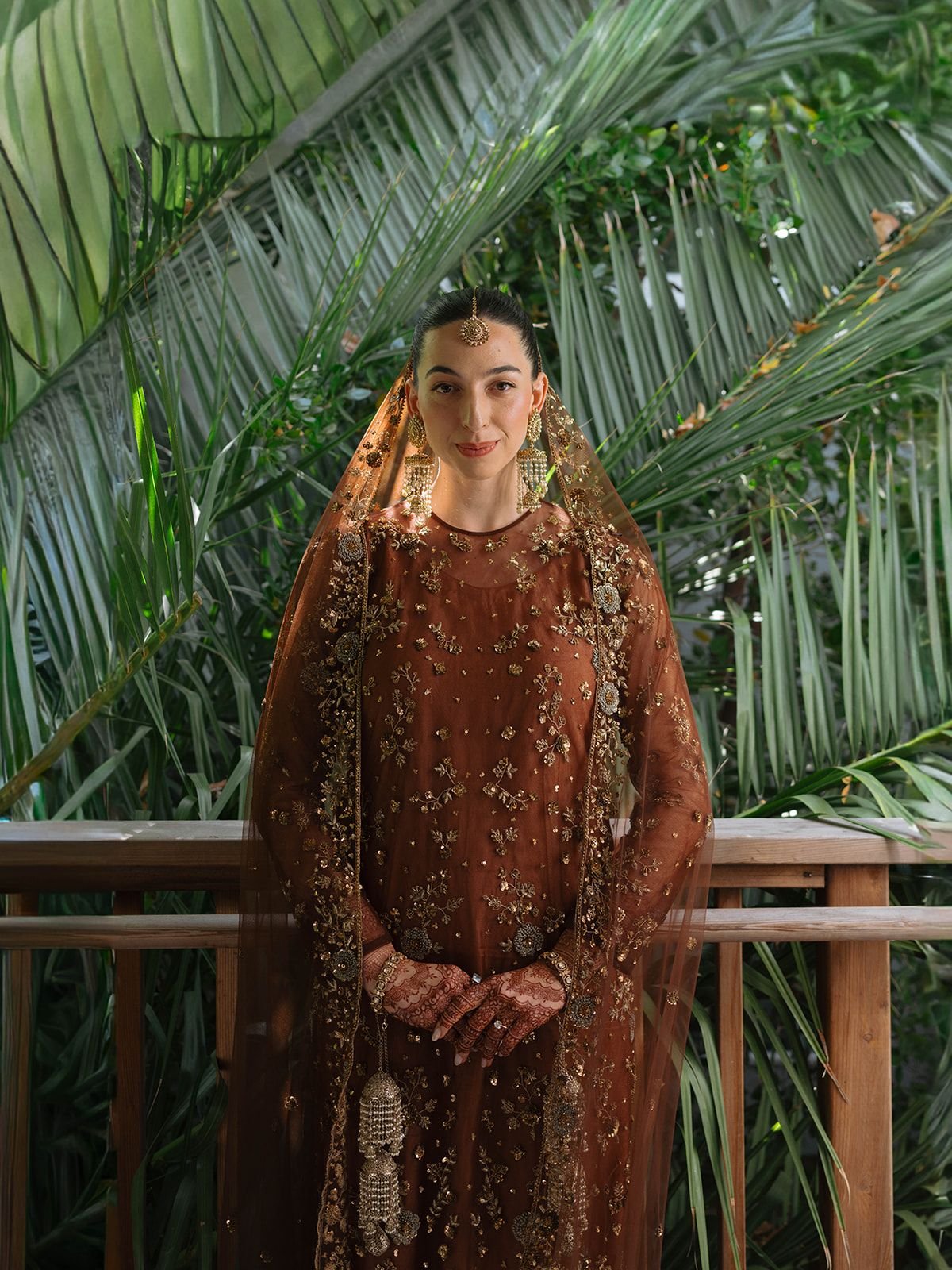 A woman dressed in traditional Indian attire, including a brown embroidered dress, a sheer embellished dupatta, and gold jewelry including earrings, a maang tikka, and bangles, stands in front of lush green tropical plants.
