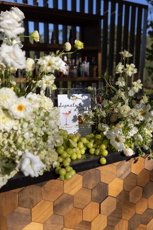 A bar decorated with white flowers and green grapes, with a sign that says 'Signature drinks' and a drawing of a cocktail glass, in an outdoor setting.