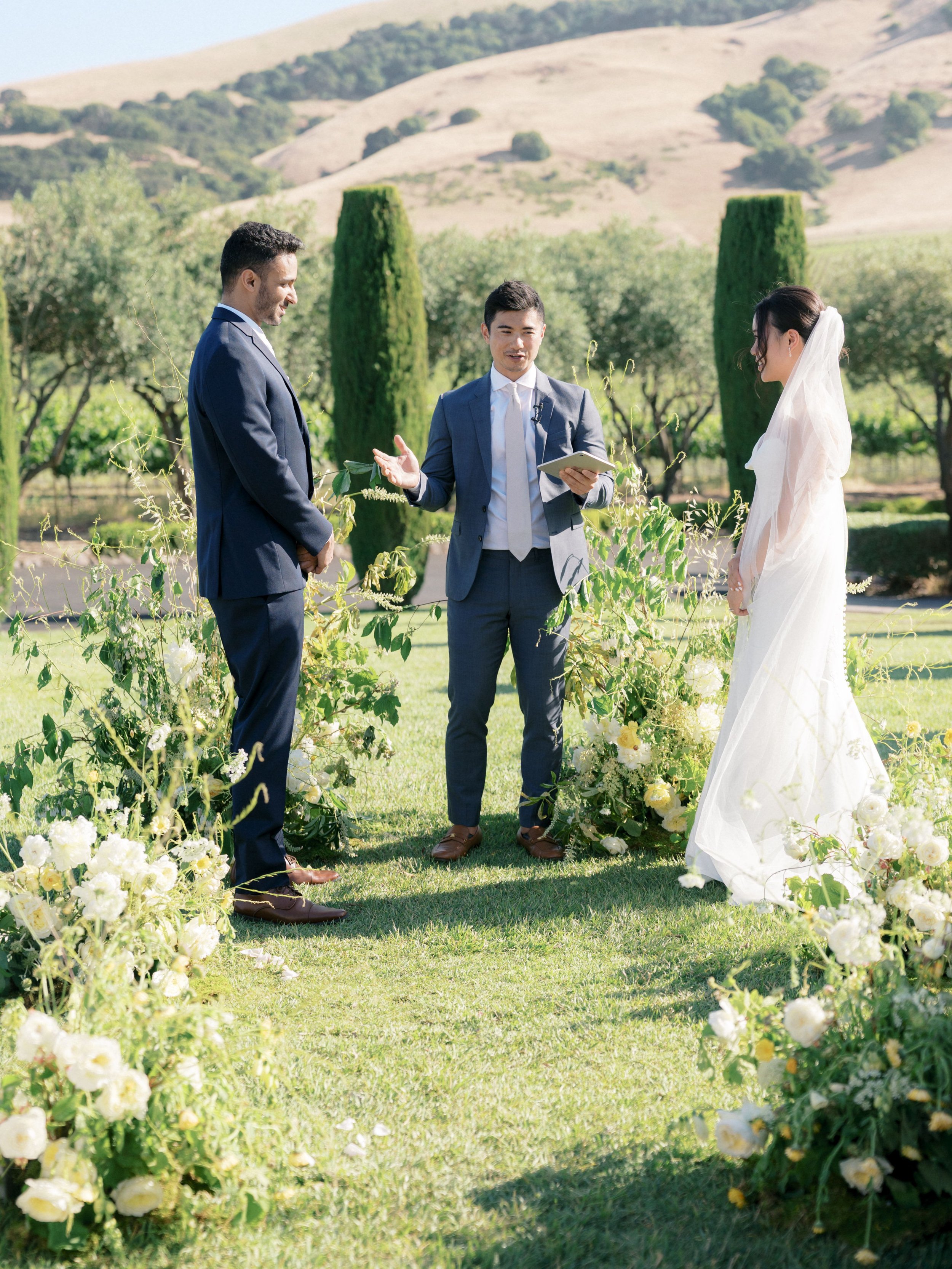 A wedding ceremony outdoors with a bride, groom, and officiant against a backdrop of grassy hills and cypress trees.