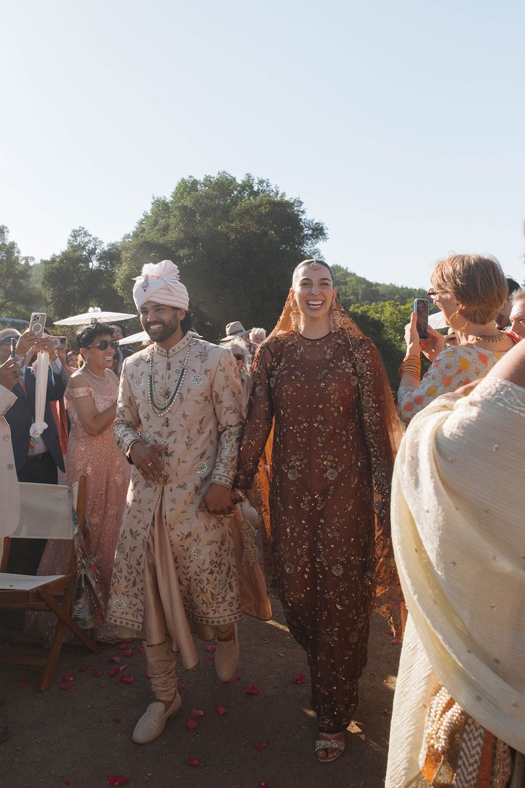 Two people in traditional Indian attire holding hands and smiling, surrounded by guests at an outdoor celebration, with a clear sky and trees in the background.