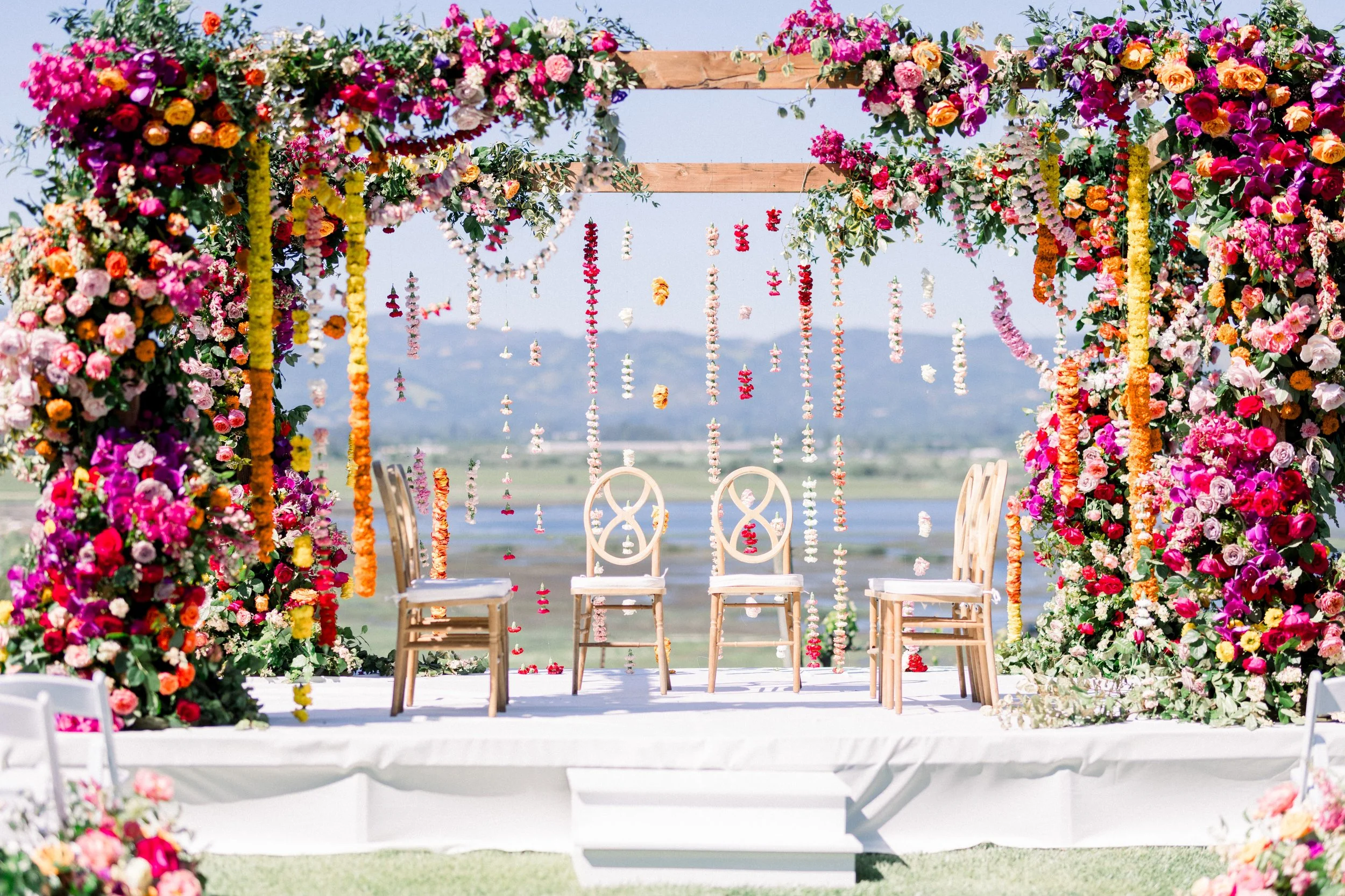 Outdoor wedding ceremony setup with a floral arch, hanging flower garlands, and white chairs, with a scenic mountain view in the background.
