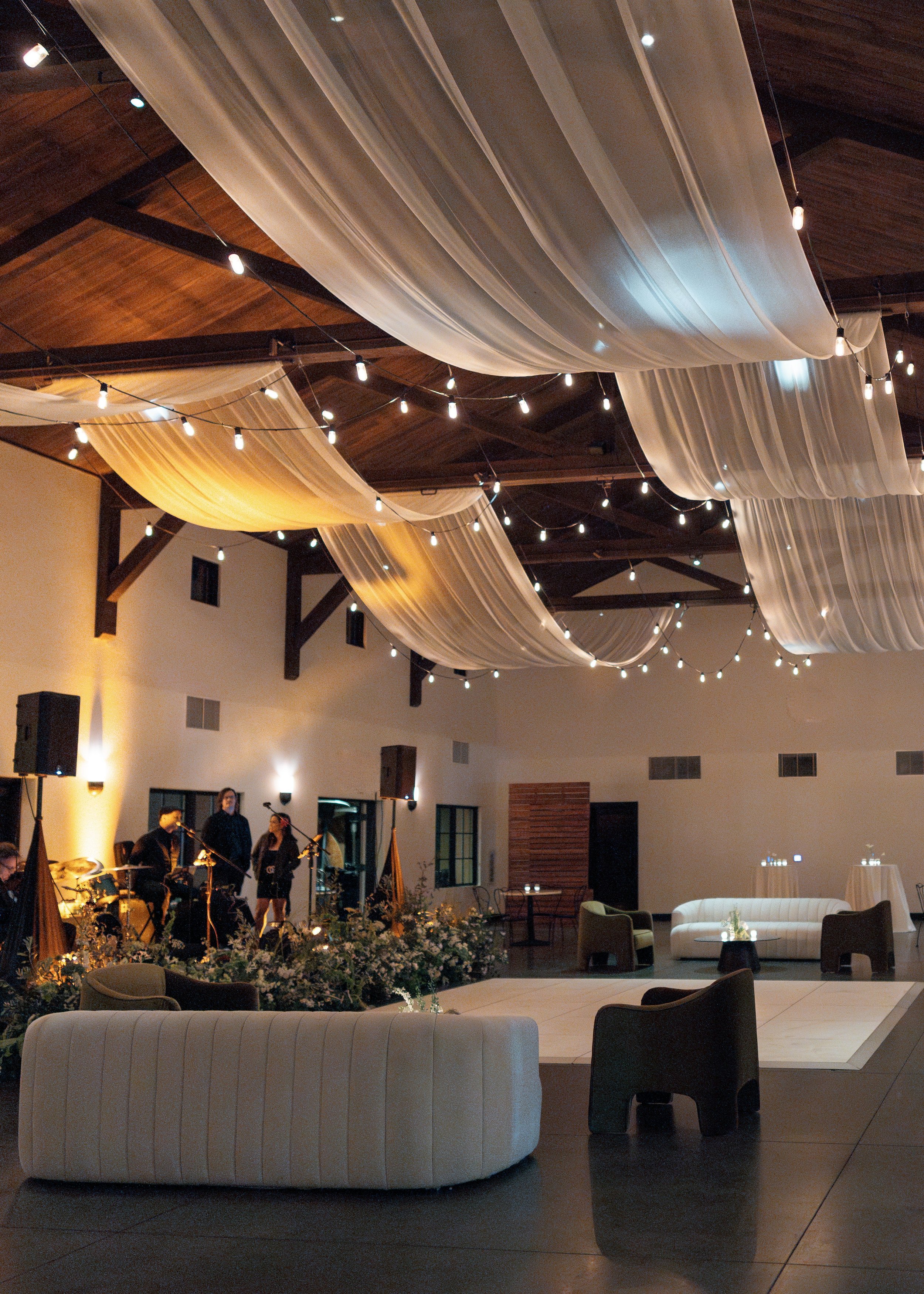 Indoor event space decorated with sheer fabric drapes hanging from the ceiling, string lights, and a stage with musicians and a speaker. The area includes lounge furniture such as sofas and chairs, with warm lighting creating a cozy ambiance.
