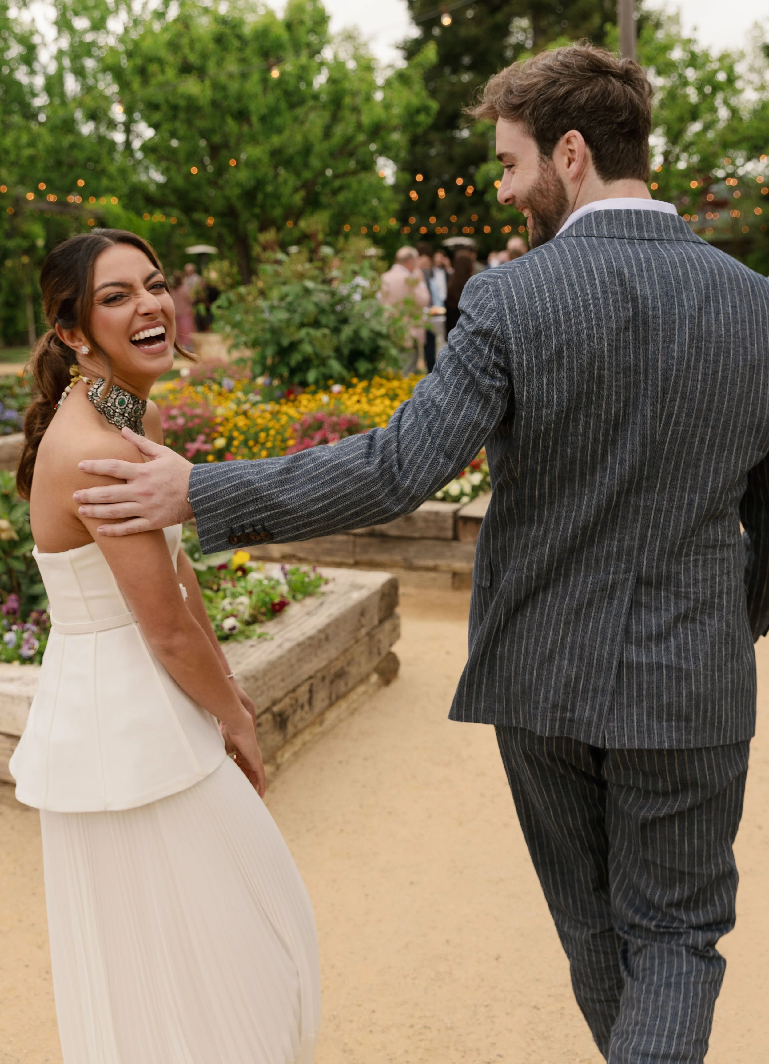 A woman in a strapless white dress laughing with a man in a dark pinstriped suit outdoors at a garden party with string lights, plants, and other guests in the background.